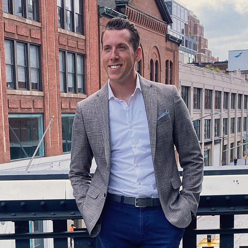 Man in blazer smiles outdoors, standing on a bridge, city buildings in the background.