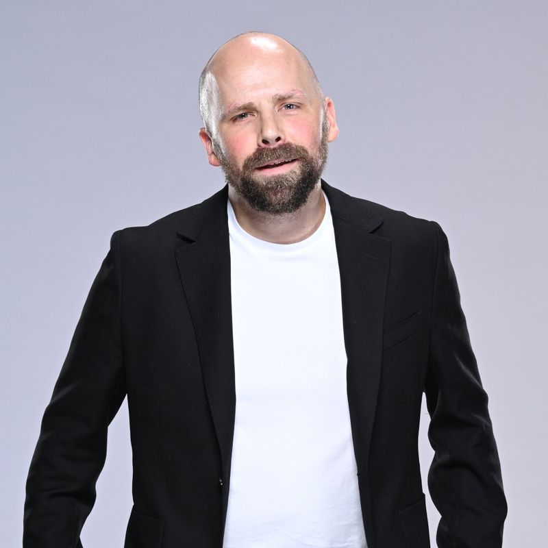 Bald man with beard wearing black blazer and white t-shirt, looking at the camera. Neutral background.