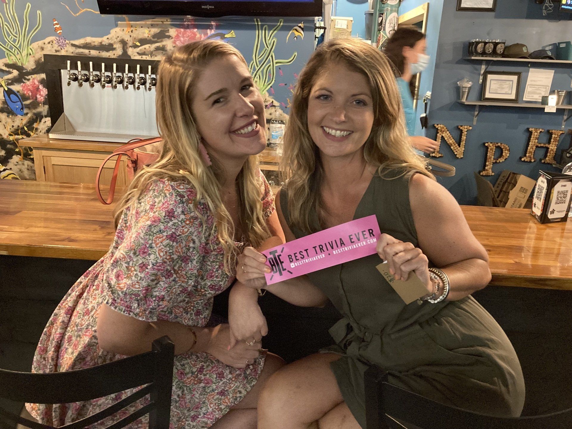 Two women smile, holding a pink sign in a bar. One wears a floral dress; the other, an olive jumpsuit.
