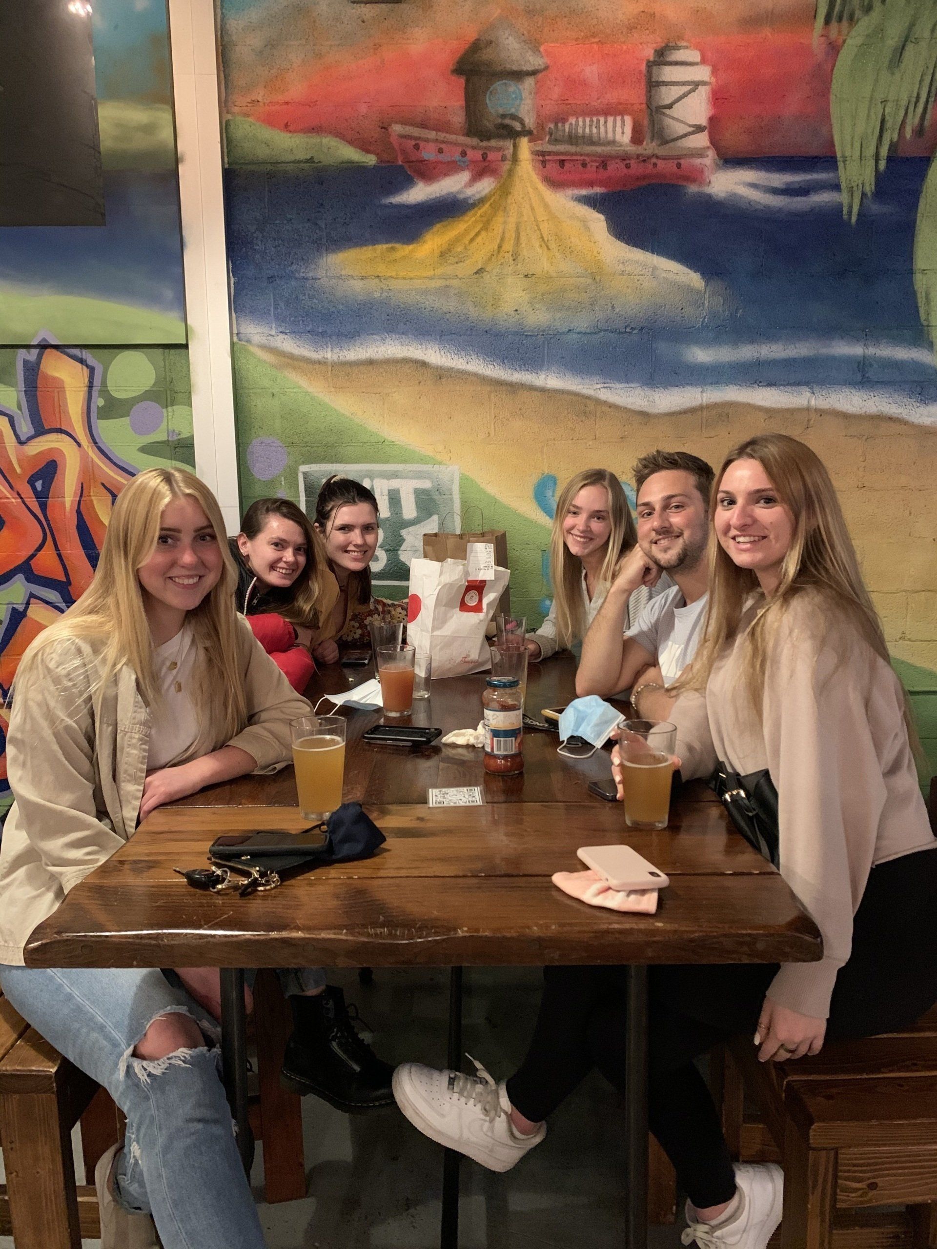 Group of people at a wooden table in a restaurant. Colorful mural background. Smiling faces. Drinks and food on table.