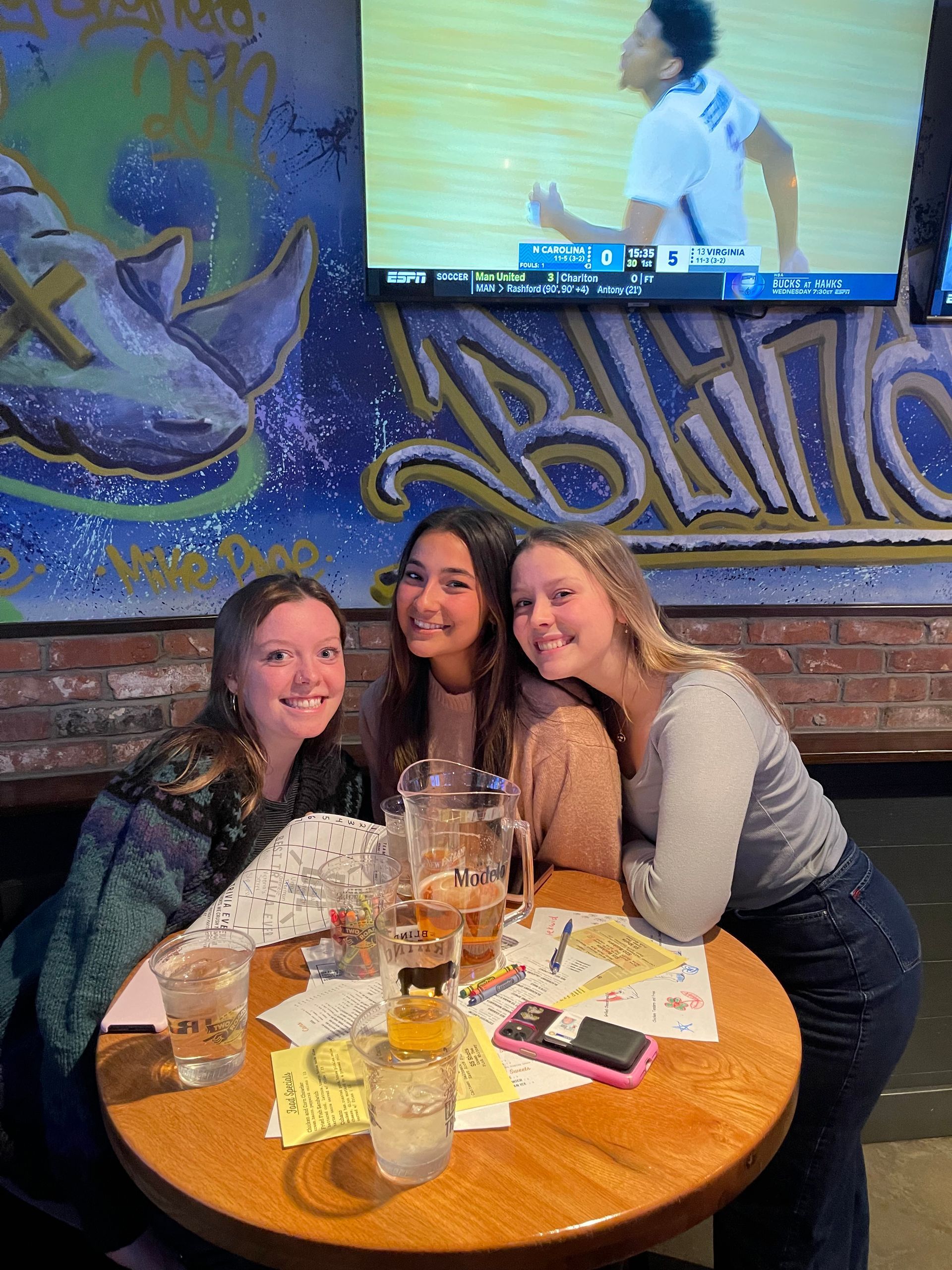 Three young people smiling around a table at a bar, drinks, a phone, and a TV showing a sports game.