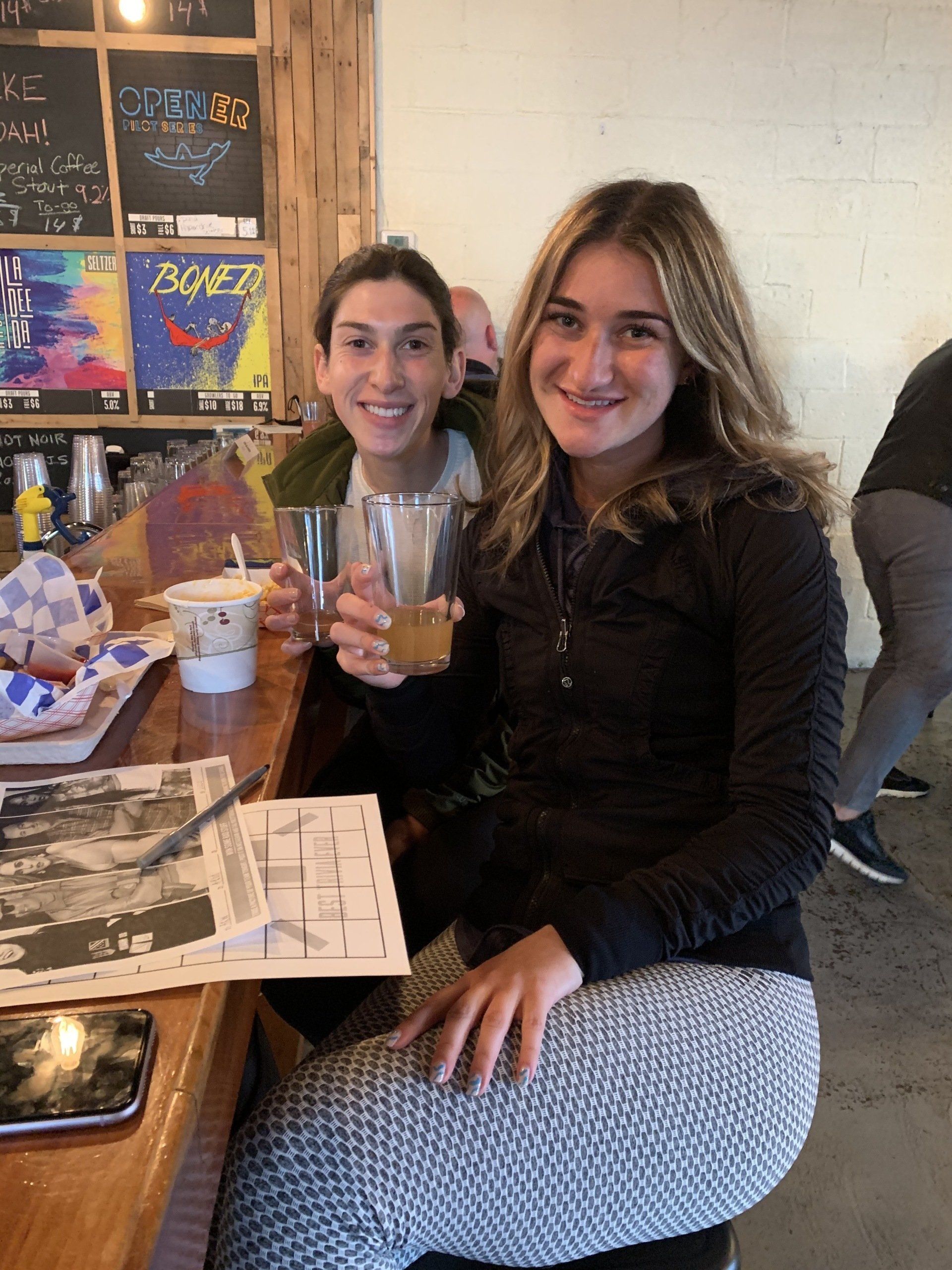 Two smiling people at a bar, holding drinks. One wears a black jacket, the other a green shirt.