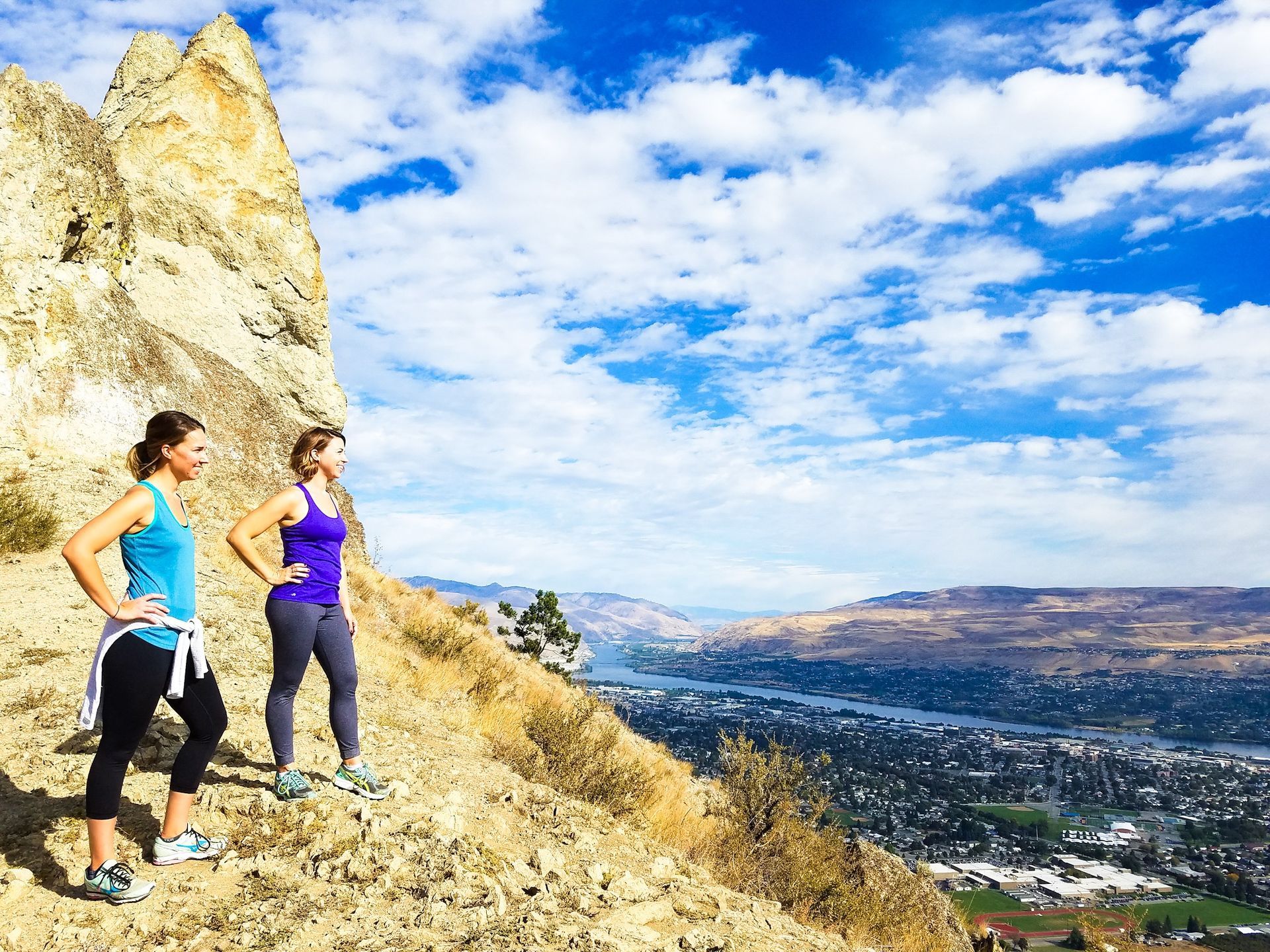 Two women are standing on top of a mountain looking out over a city.