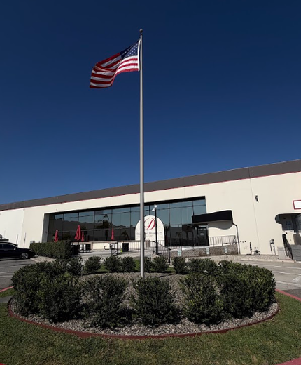 American flag on a pole in front of a building with a landscaped shrub bed under a clear blue sky
