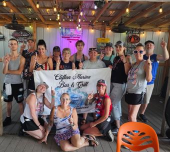 Group posing with a banner at a bar, with neon lights and people in casual party attire.