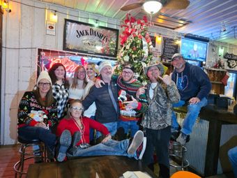 Group of people posing indoors in holiday sweaters and Santa hats beside a decorated Christmas tree.