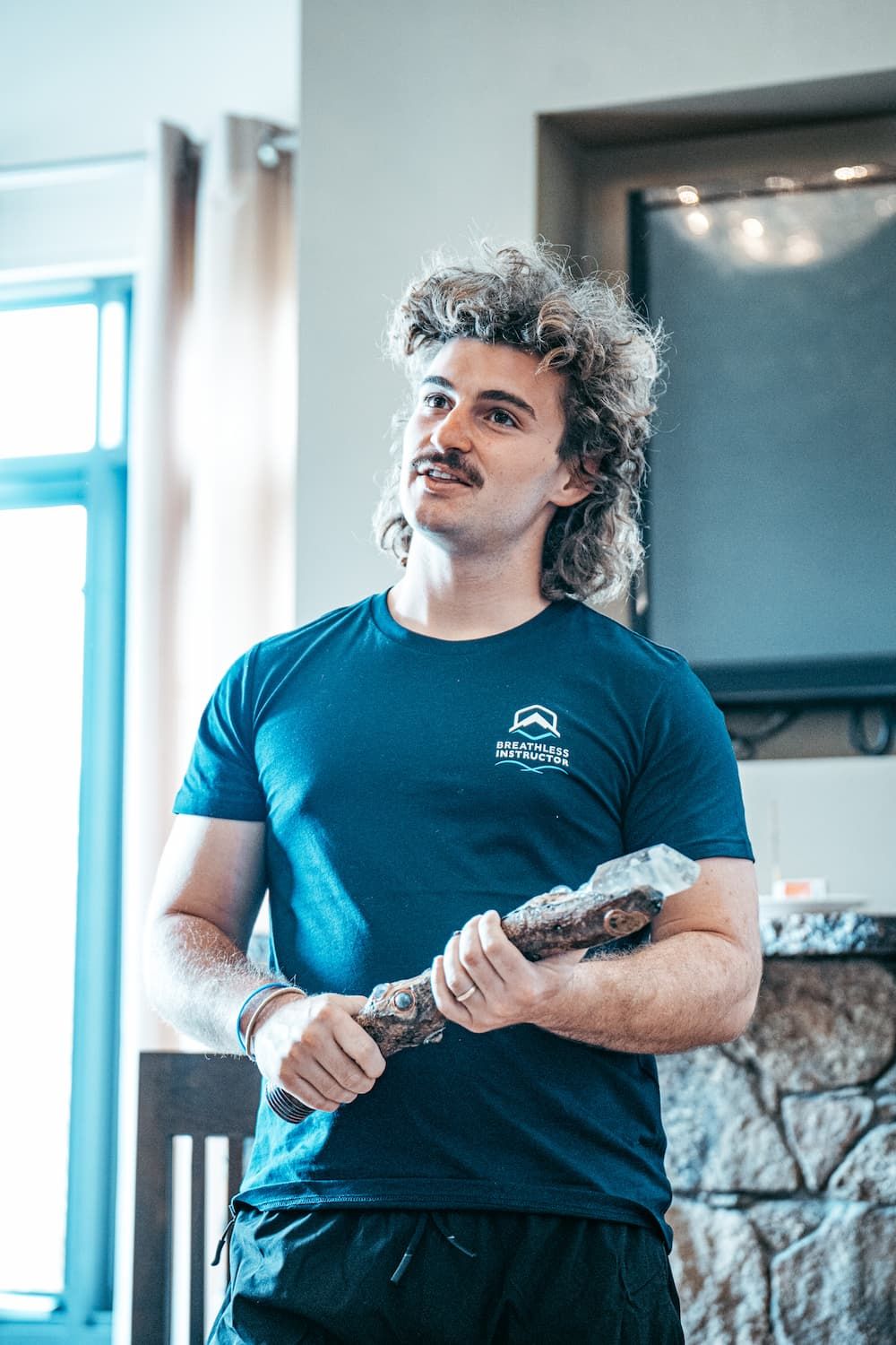 A man with curly hair and a mustache is holding a stick in a living room.