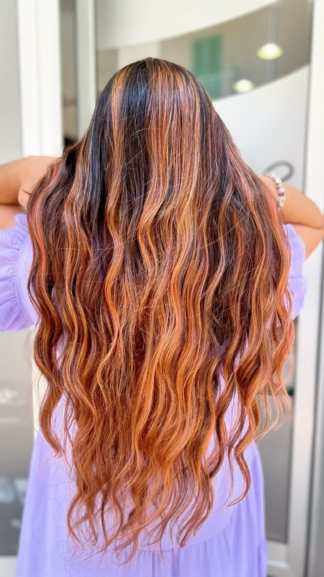 Una donna con un vestito viola e lunghi capelli ondulati è in piedi davanti a uno specchio.