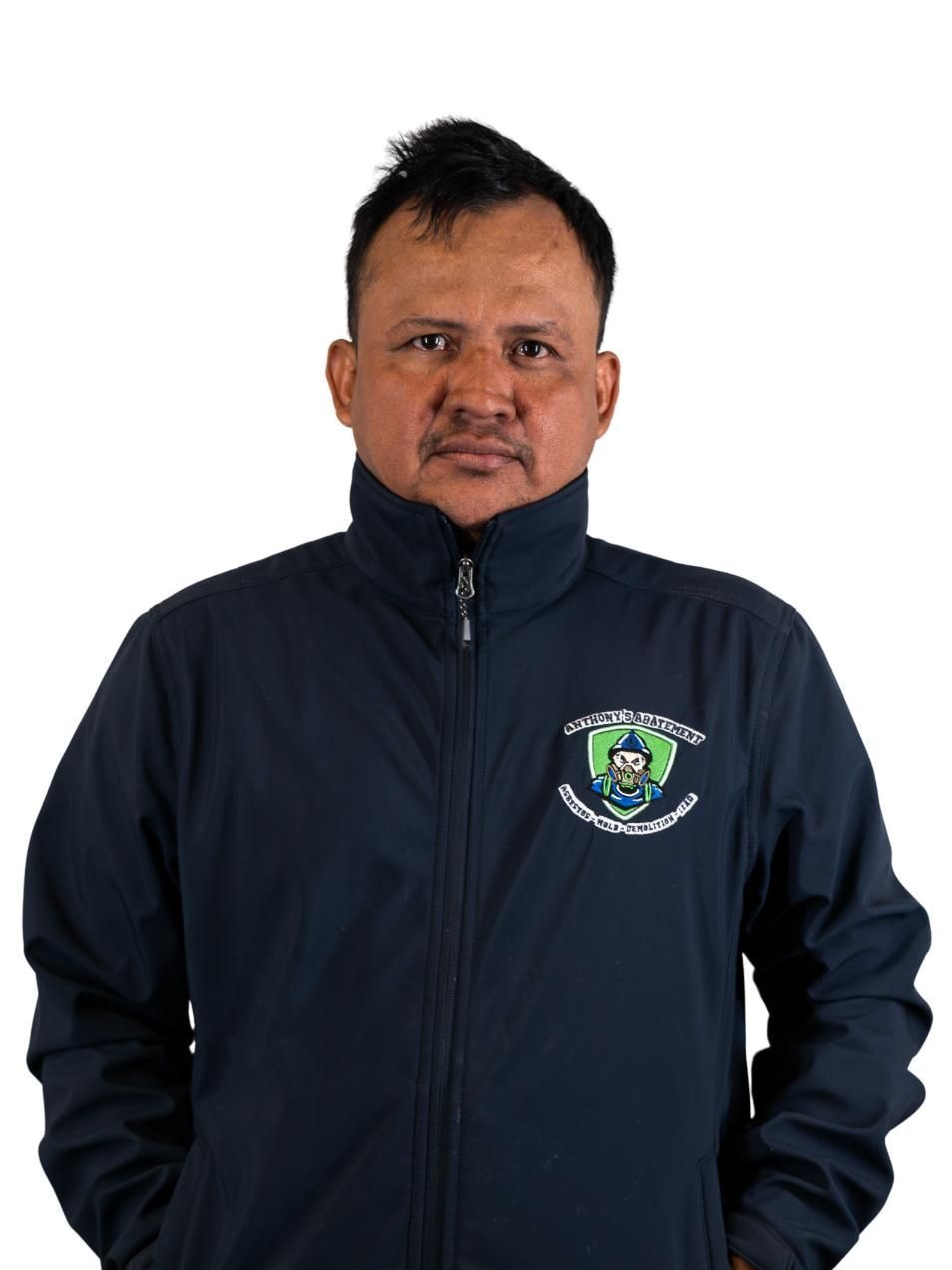 Man in dark jacket with logo, standing against a white background, hands in pockets.