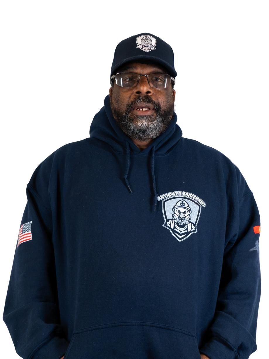 Man in navy hoodie and cap with firefighter-style emblem, standing against a white background.
