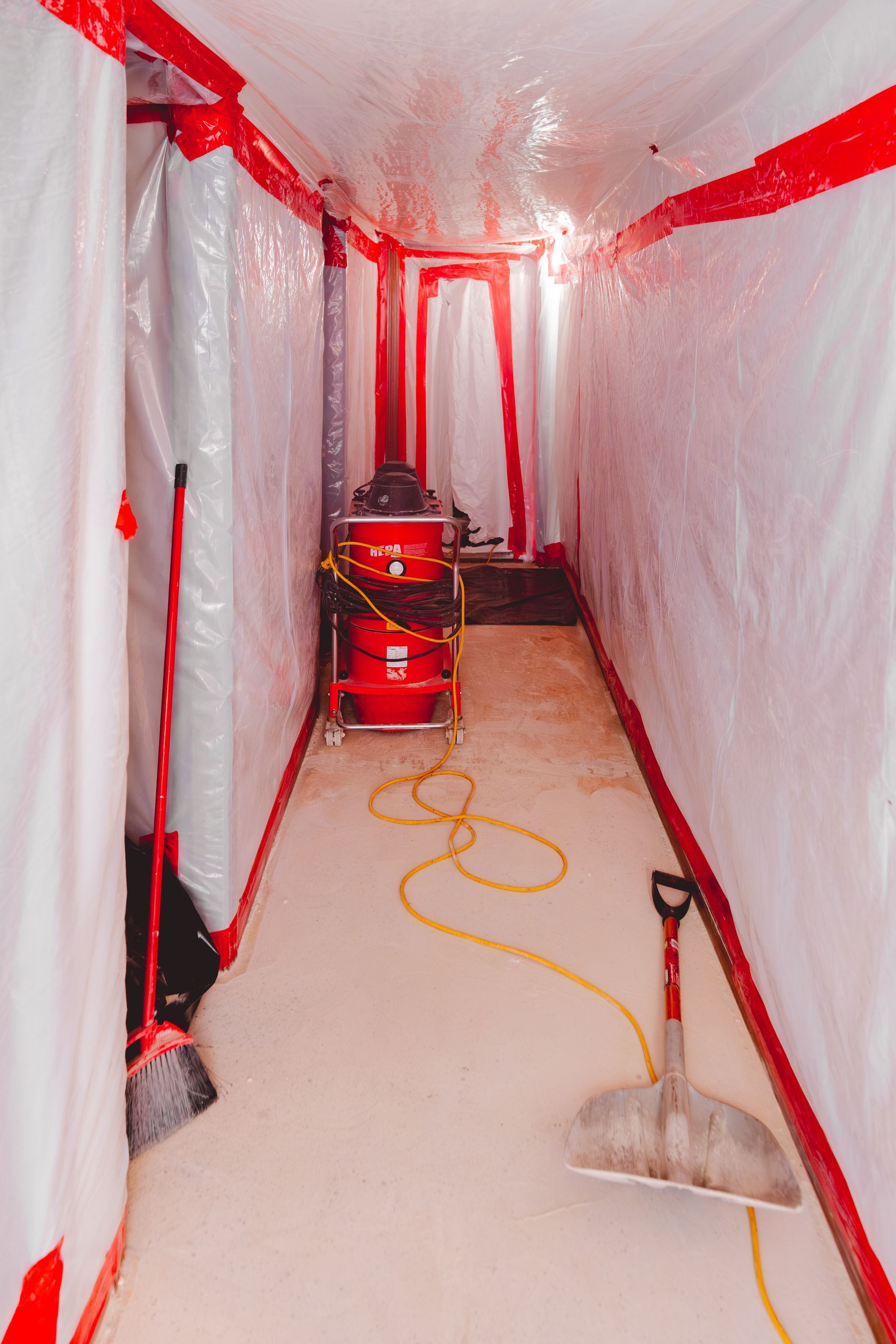 Narrow plastic-covered hallway with red tape, a vacuum, hose, and a dustpan on the floor