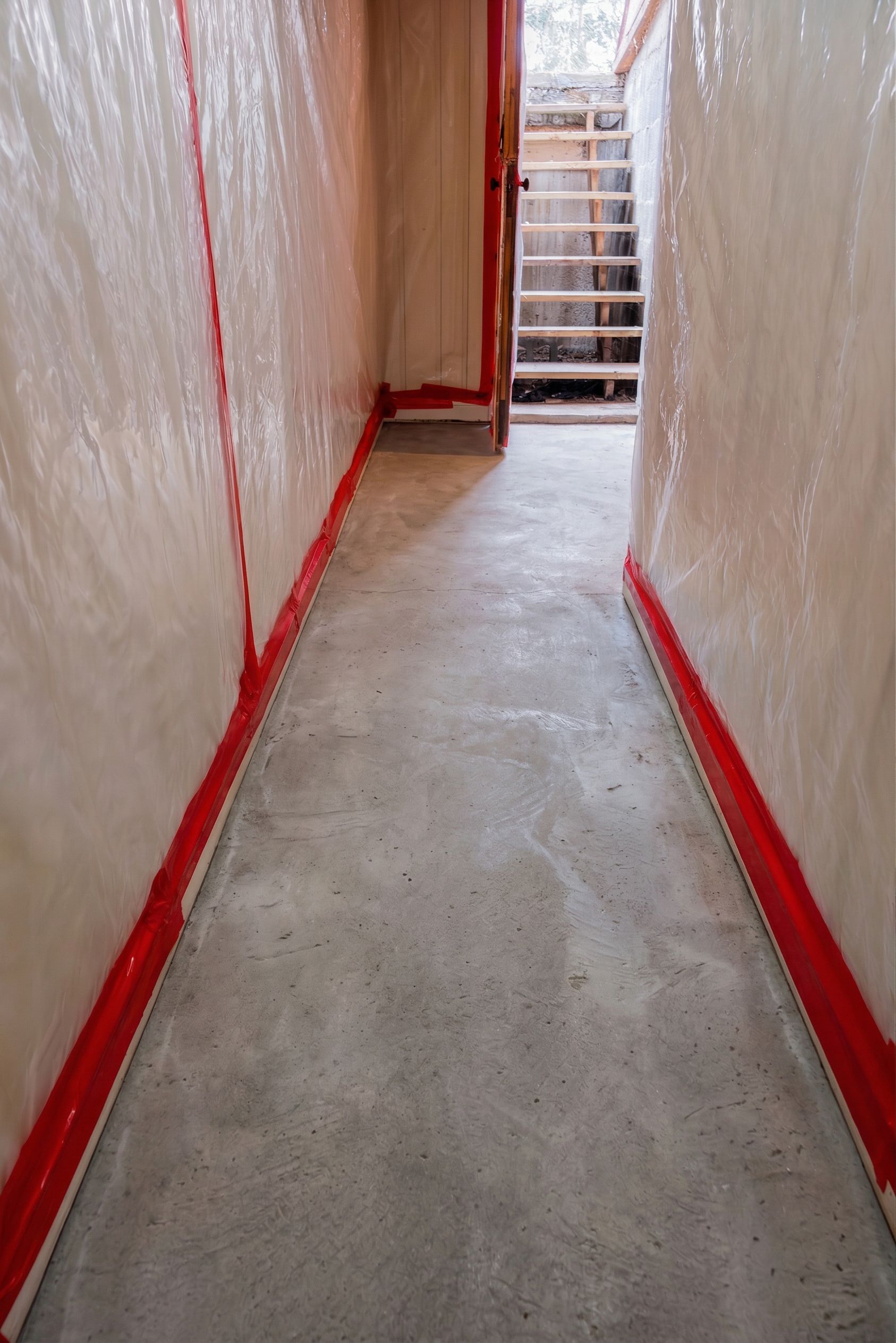 Narrow unfinished hallway with plastic-covered walls and red tape leading to a stairway ahead