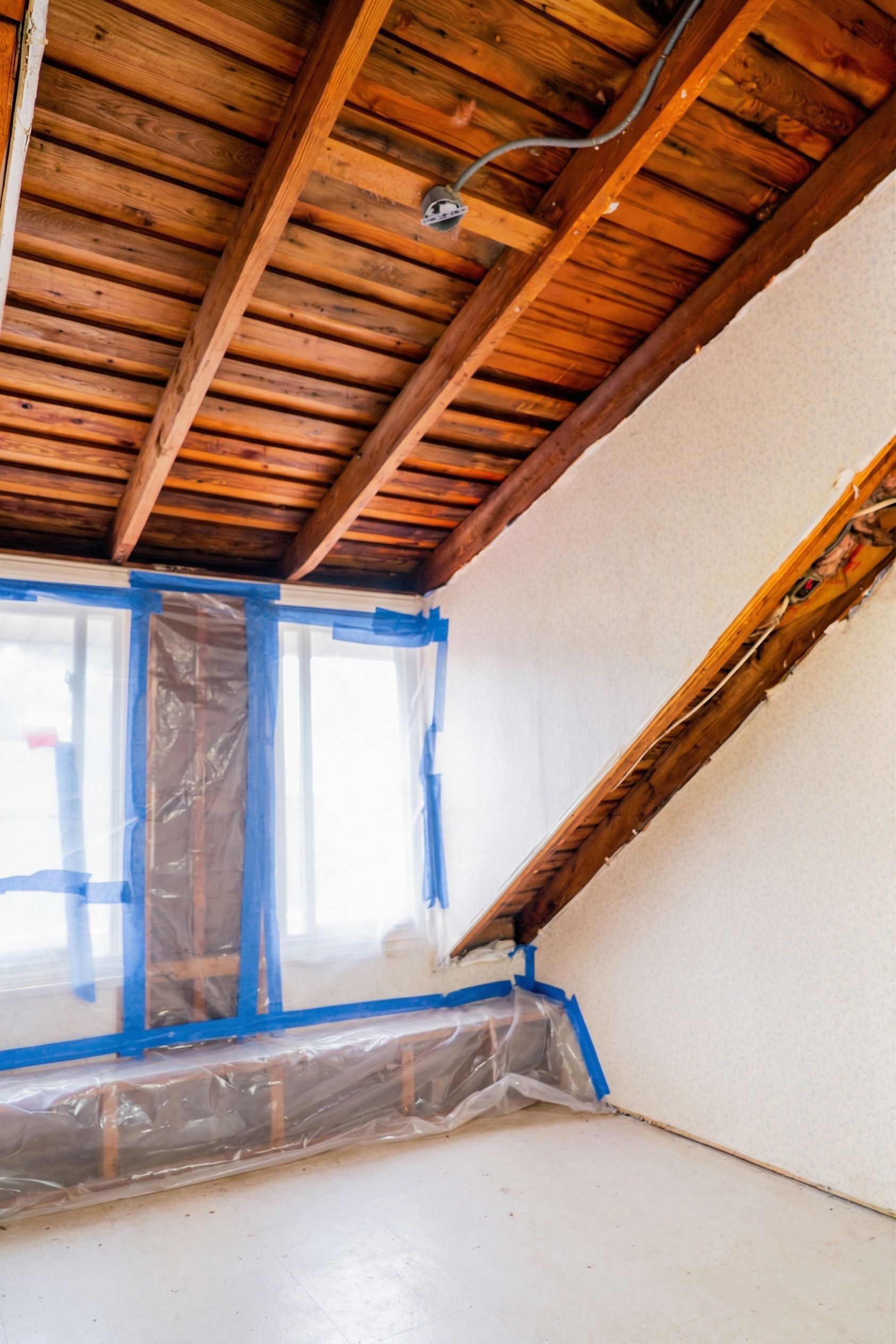 Attic room with exposed wooden ceiling, white walls, and windows covered in blue tape and plastic sheeting