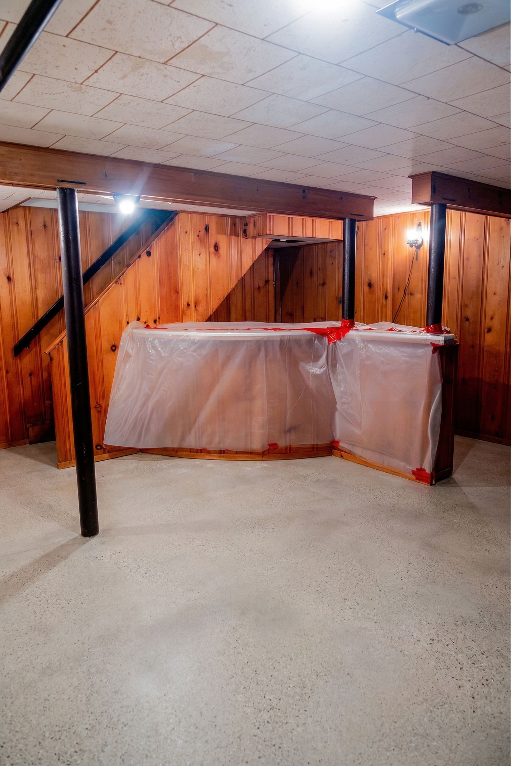 Basement room with wood-paneled walls, exposed beams, and plastic-covered furniture against the wall