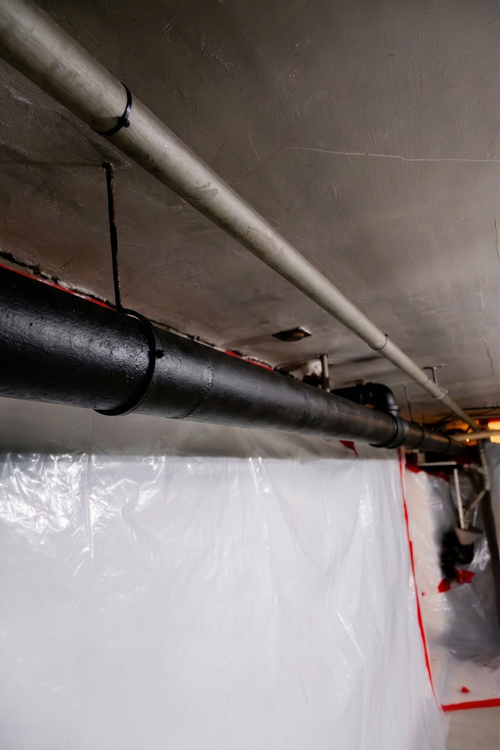 Basement ceiling with black and white pipes above plastic sheeting and red tape boundary