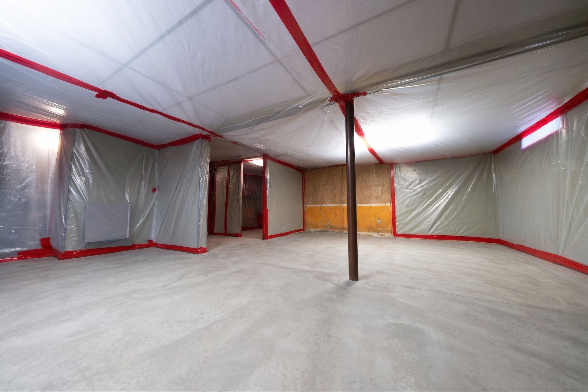 Empty unfinished room wrapped in plastic sheeting, with exposed ceiling, red tape, and a support pole.