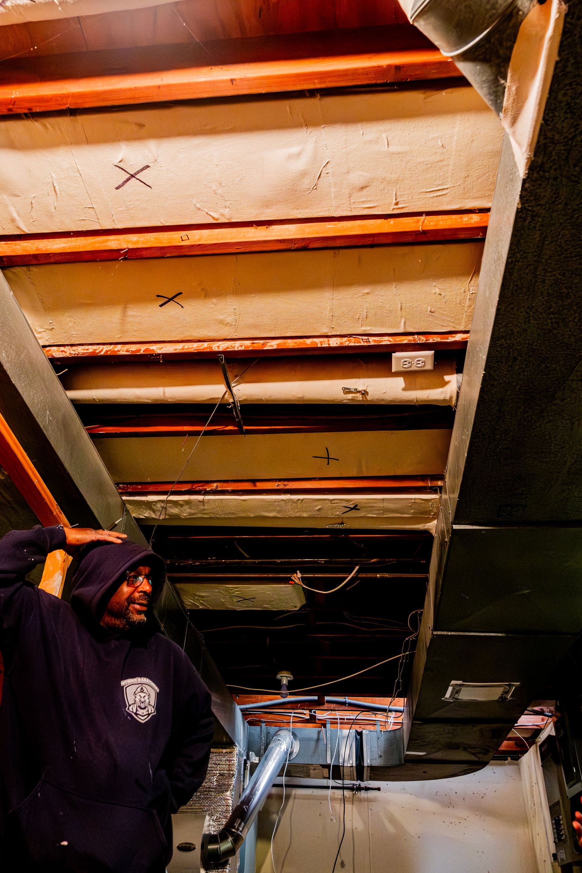 Person in a dark hoodie looking up at exposed wooden beams in a narrow basement corridor.