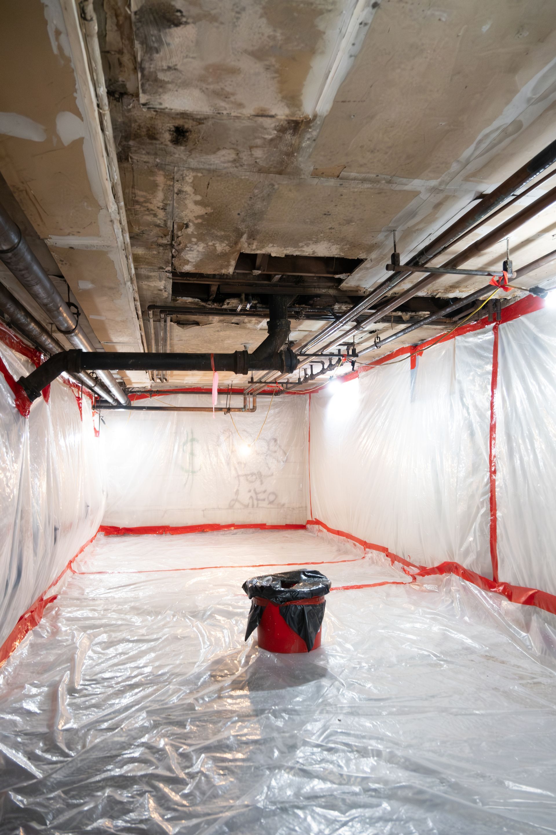 Plastic-covered room with exposed ceiling pipes and a red bucket in the center, resembling a renovation site