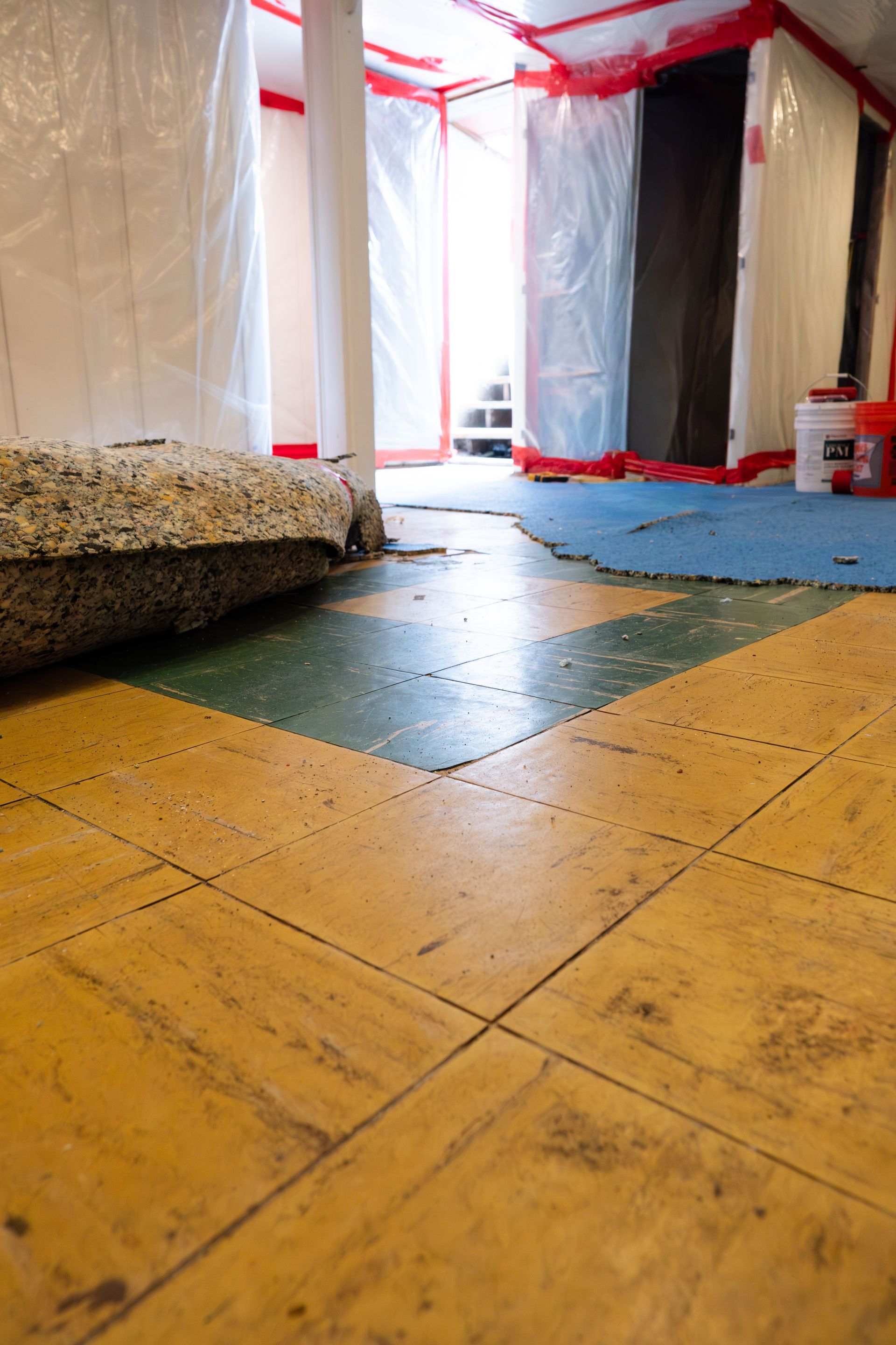 Hallway under renovation with plastic-covered walls, red tape, and yellow wood flooring leading to a doorway.