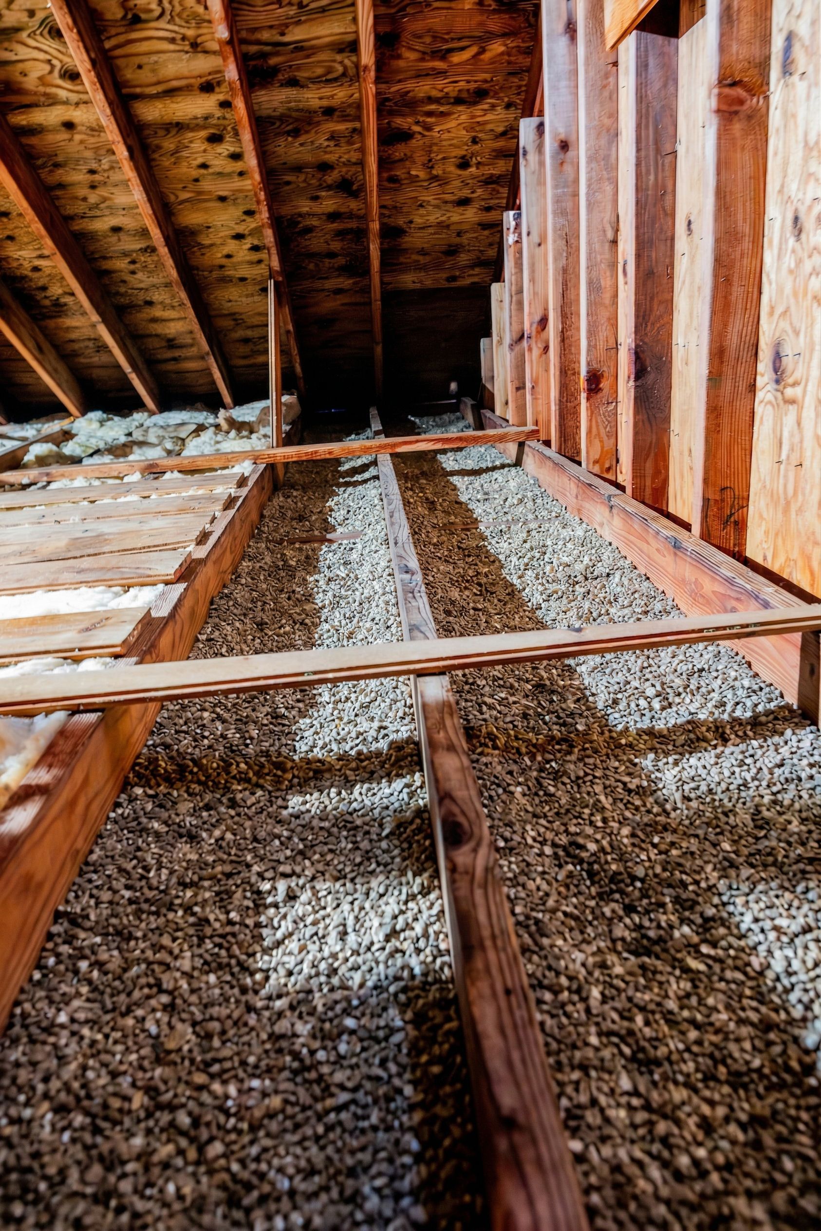 Exposed attic floor joists with gravel-filled cavities and wooden framing under a sloped roof