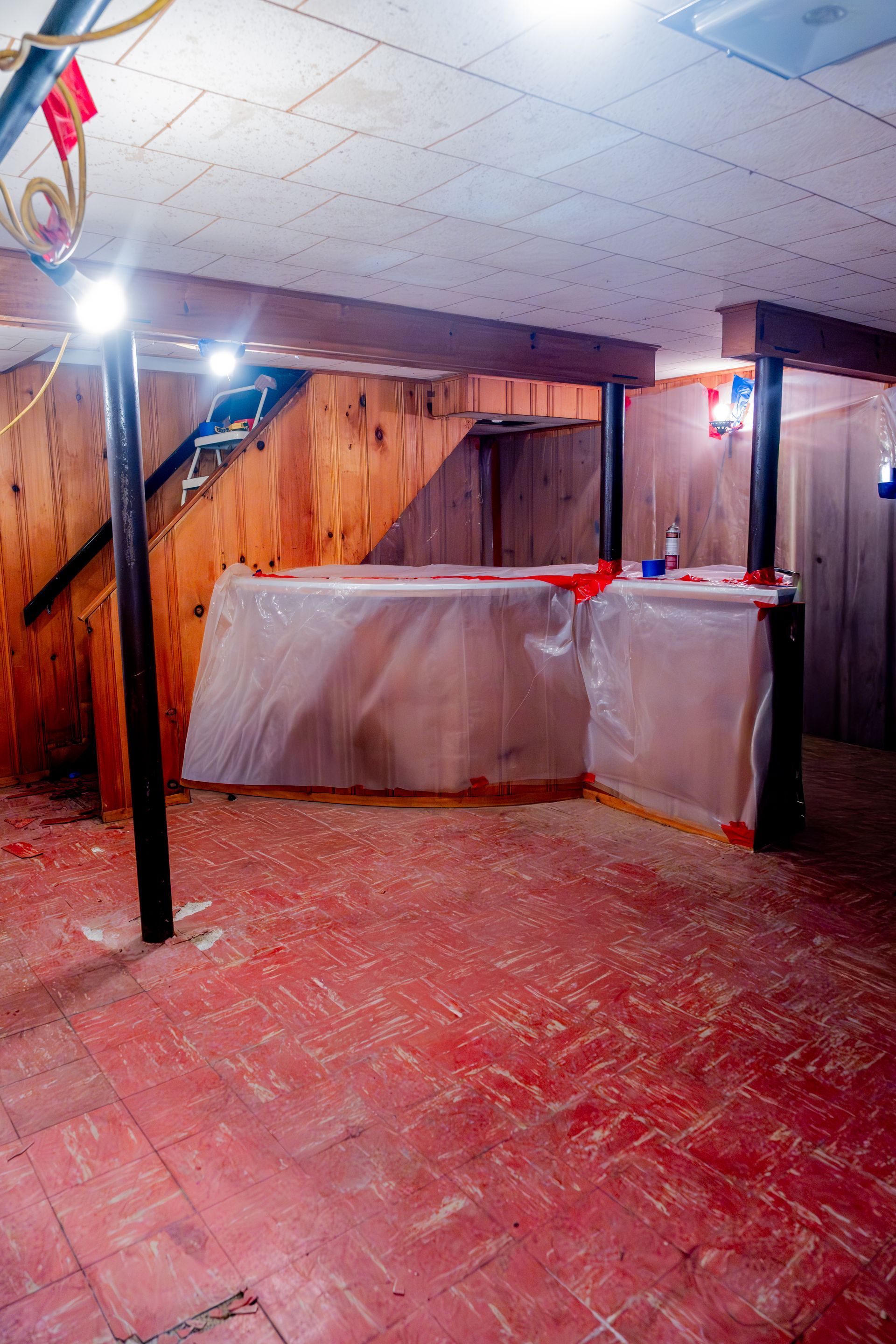 Basement room with red floor, wood-paneled walls, and plastic-covered furniture under low ceiling lights