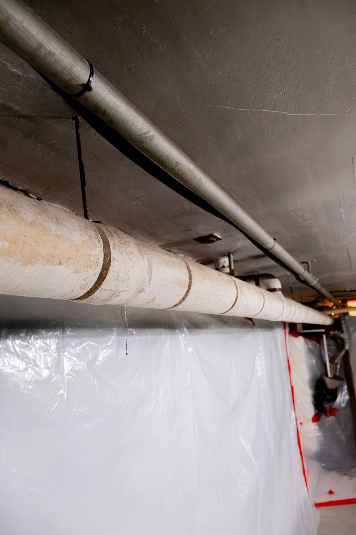 Industrial ceiling with long white pipes running above a plastic-covered wall in a narrow corridor