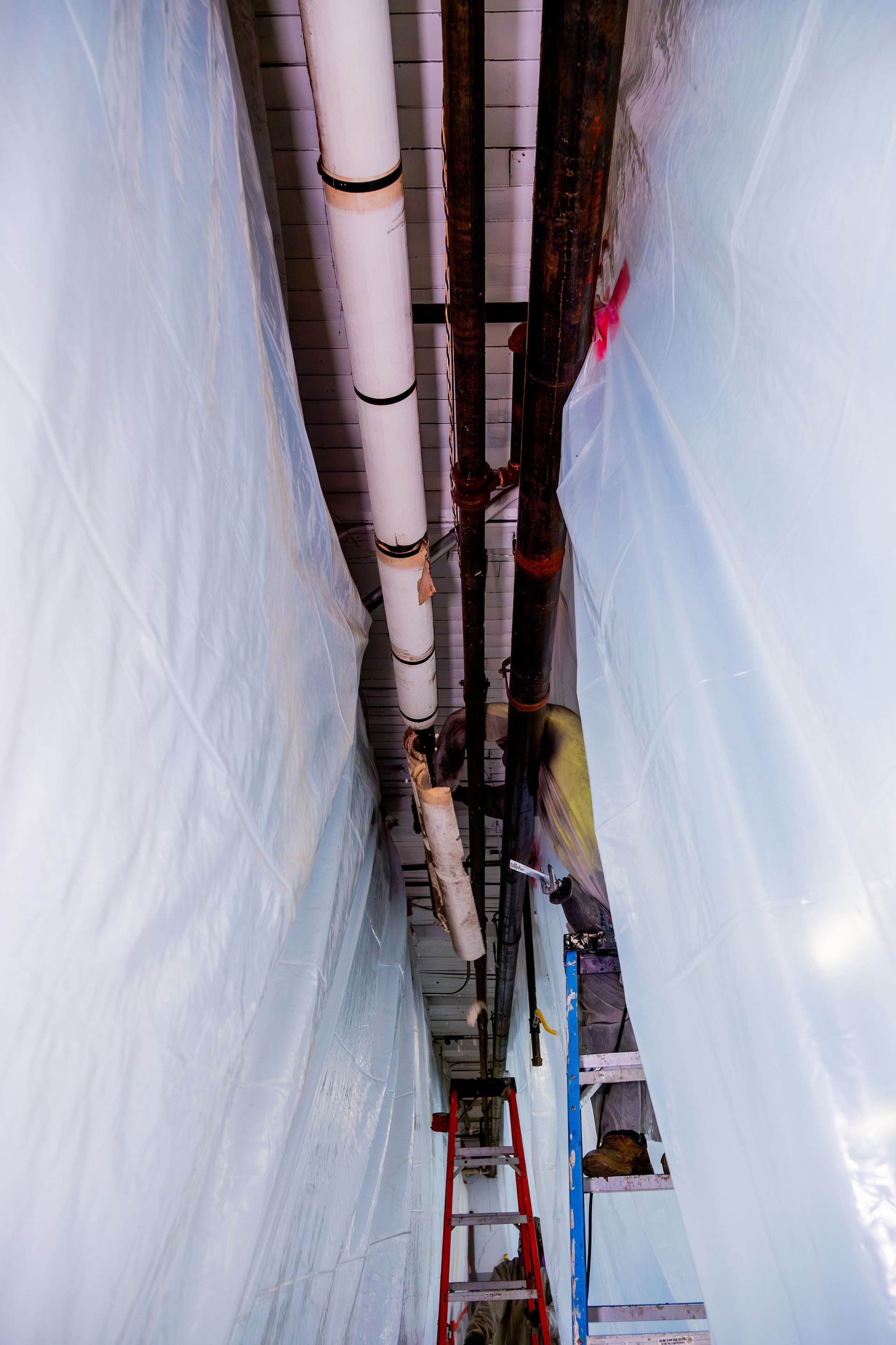 Narrow construction shaft with pipes, hoses, and a ladder between white tarp-covered walls.
