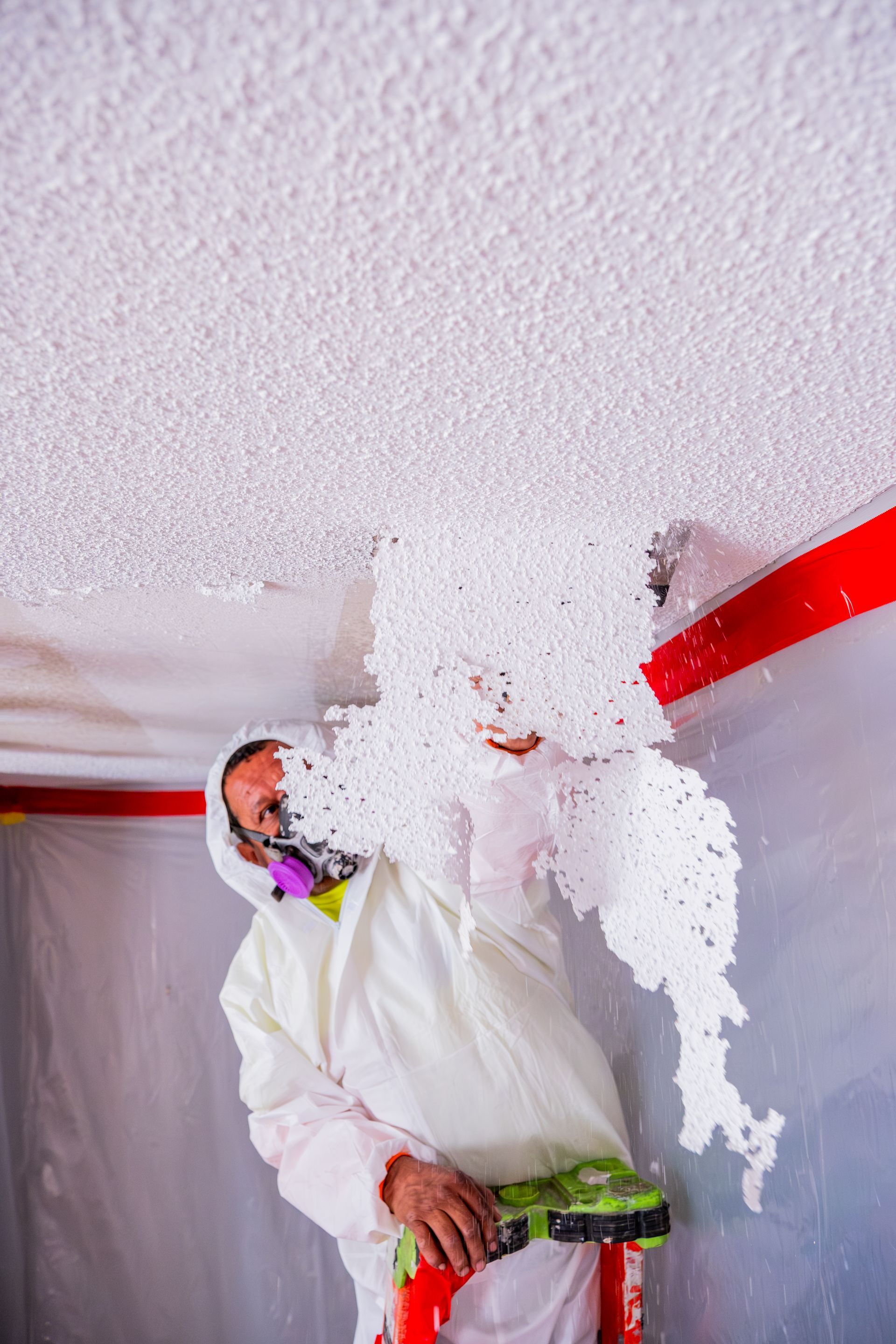 Person in white protective suit painting a ceiling, with a red roller pole and paint on the wall edge.