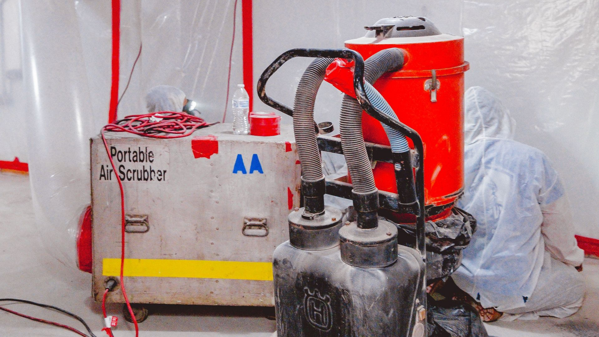Portable air scrubber and worker in hazmat suit inside a containment area, likely for asbestos abatement.