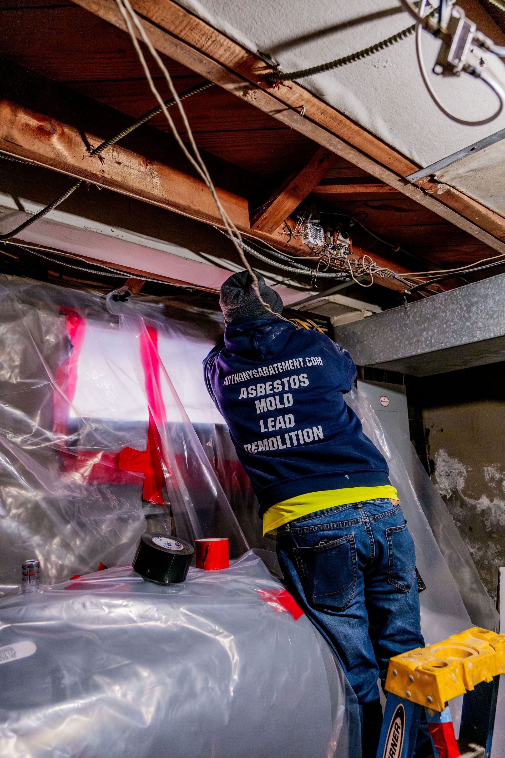 Worker in a basement crawlspace fixing exposed pipes beneath ceiling joists, covered by plastic sheeting.