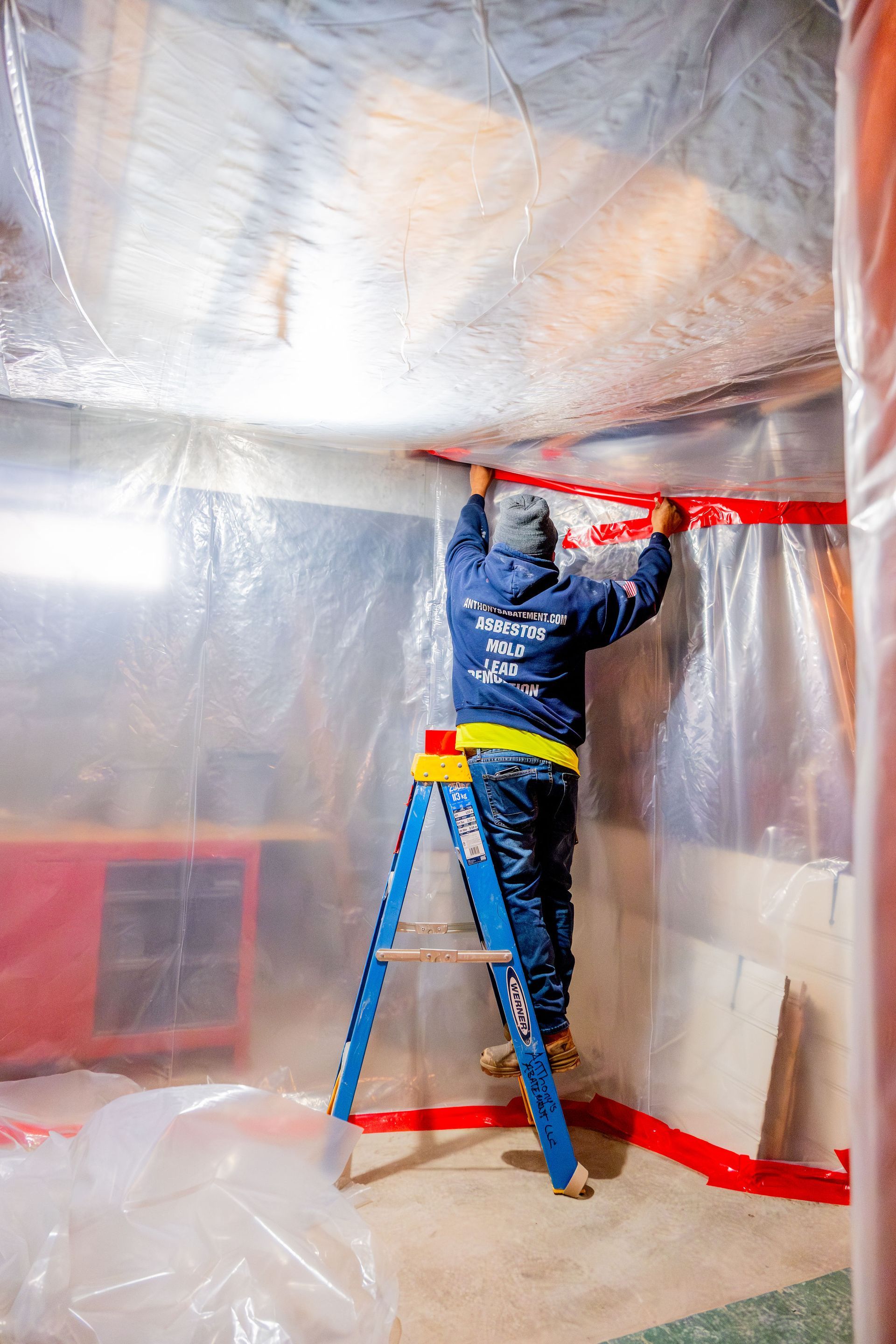 Worker on a ladder sealing a room with red tape and plastic sheeting during renovation