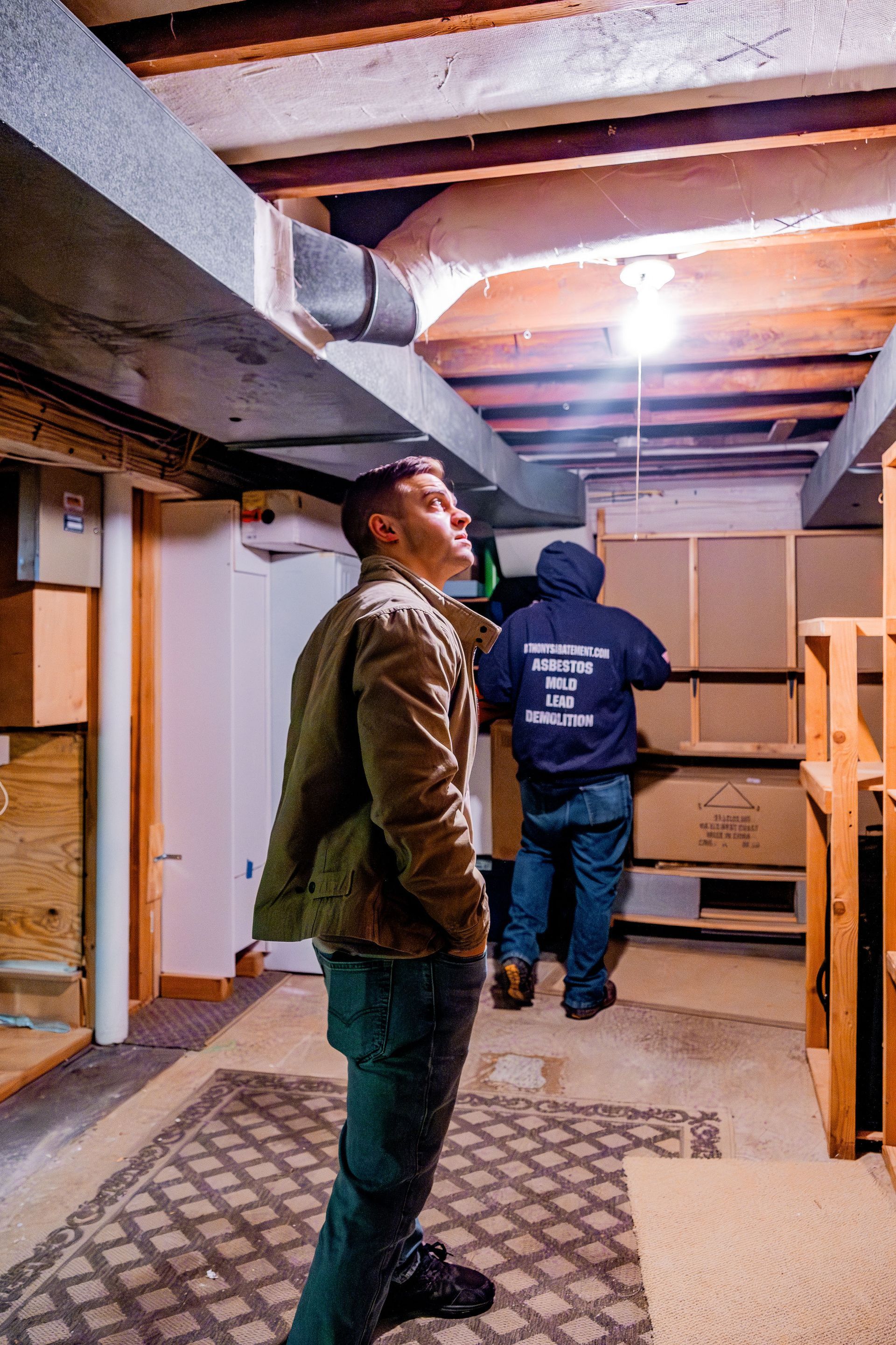 Two people standing in an unfinished basement, looking up at exposed beams and a lit ceiling bulb.