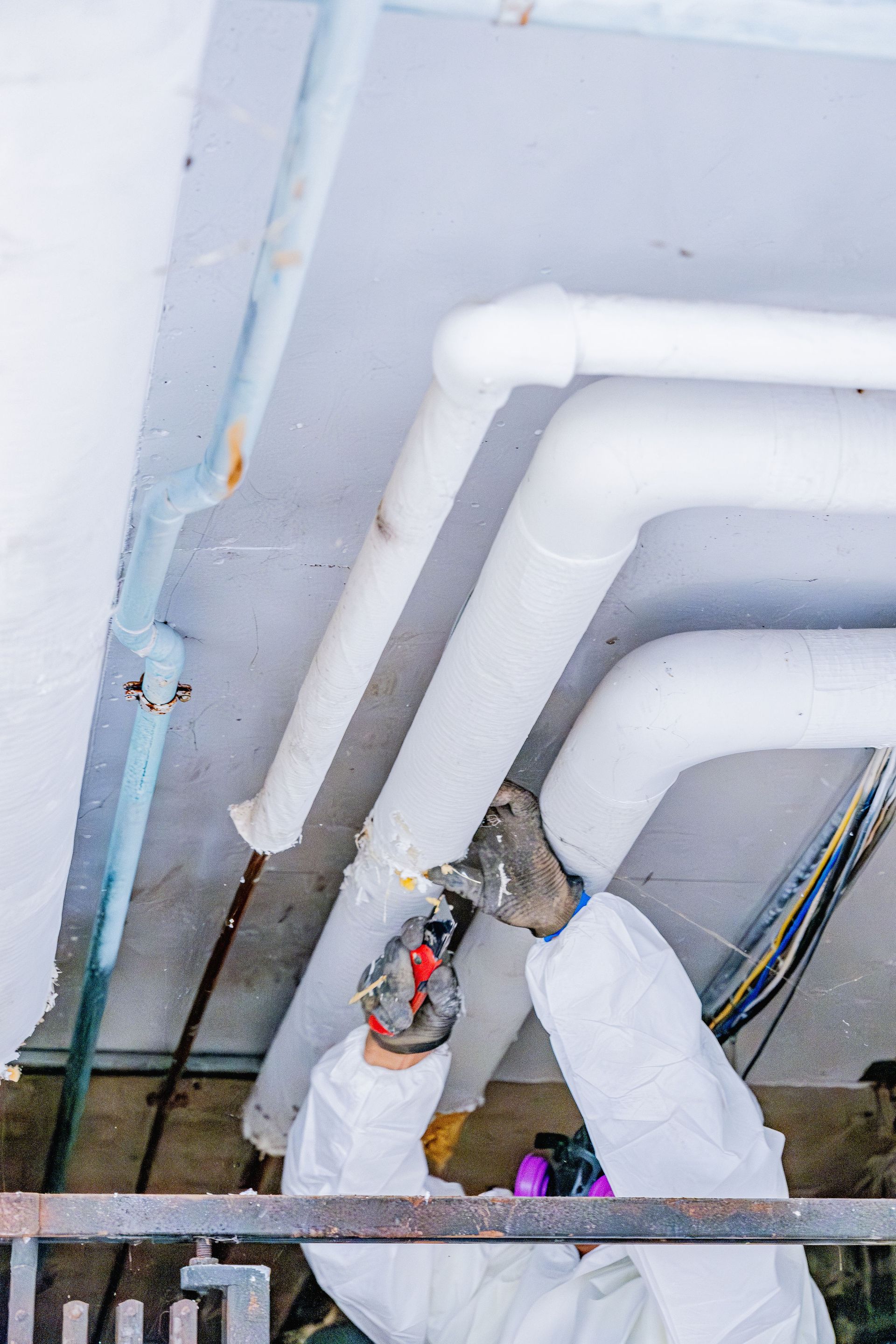 Two workers painting white ceiling pipes with sprayers in an indoor construction area