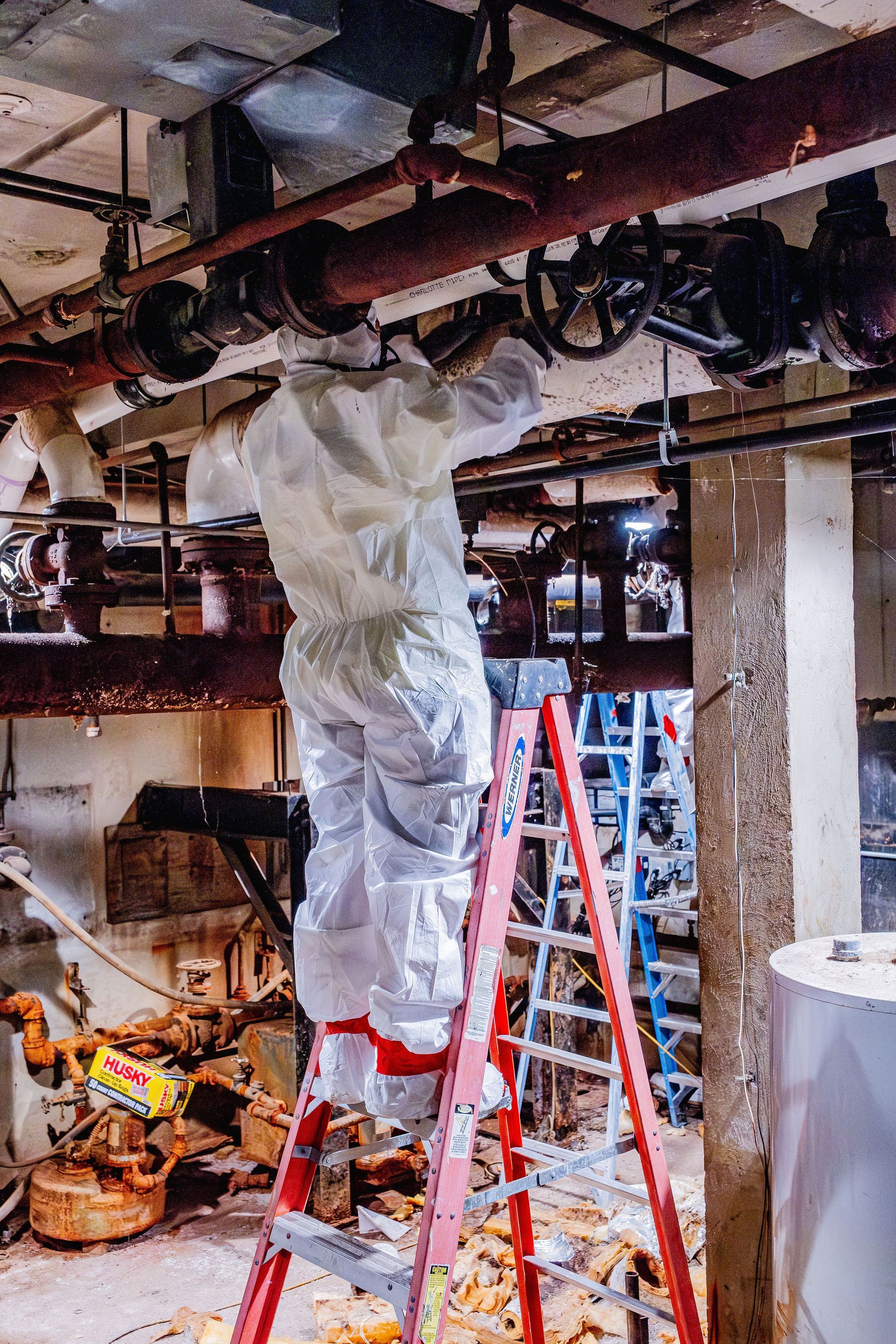 Worker on a ladder wrapping overhead pipes in plastic inside a basement utility room.