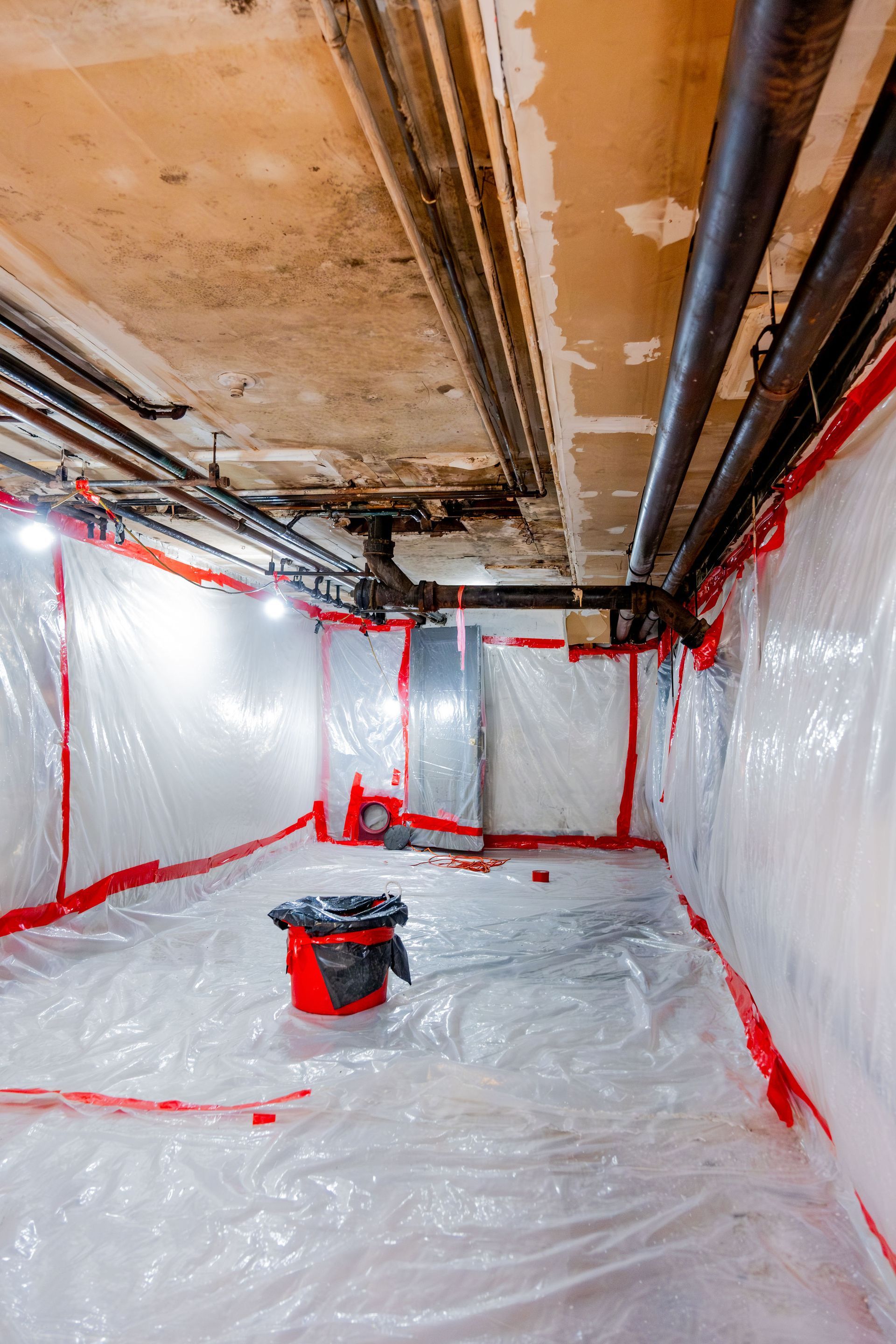 Plastic-covered basement under renovation with red tape and a paint bucket on the floor