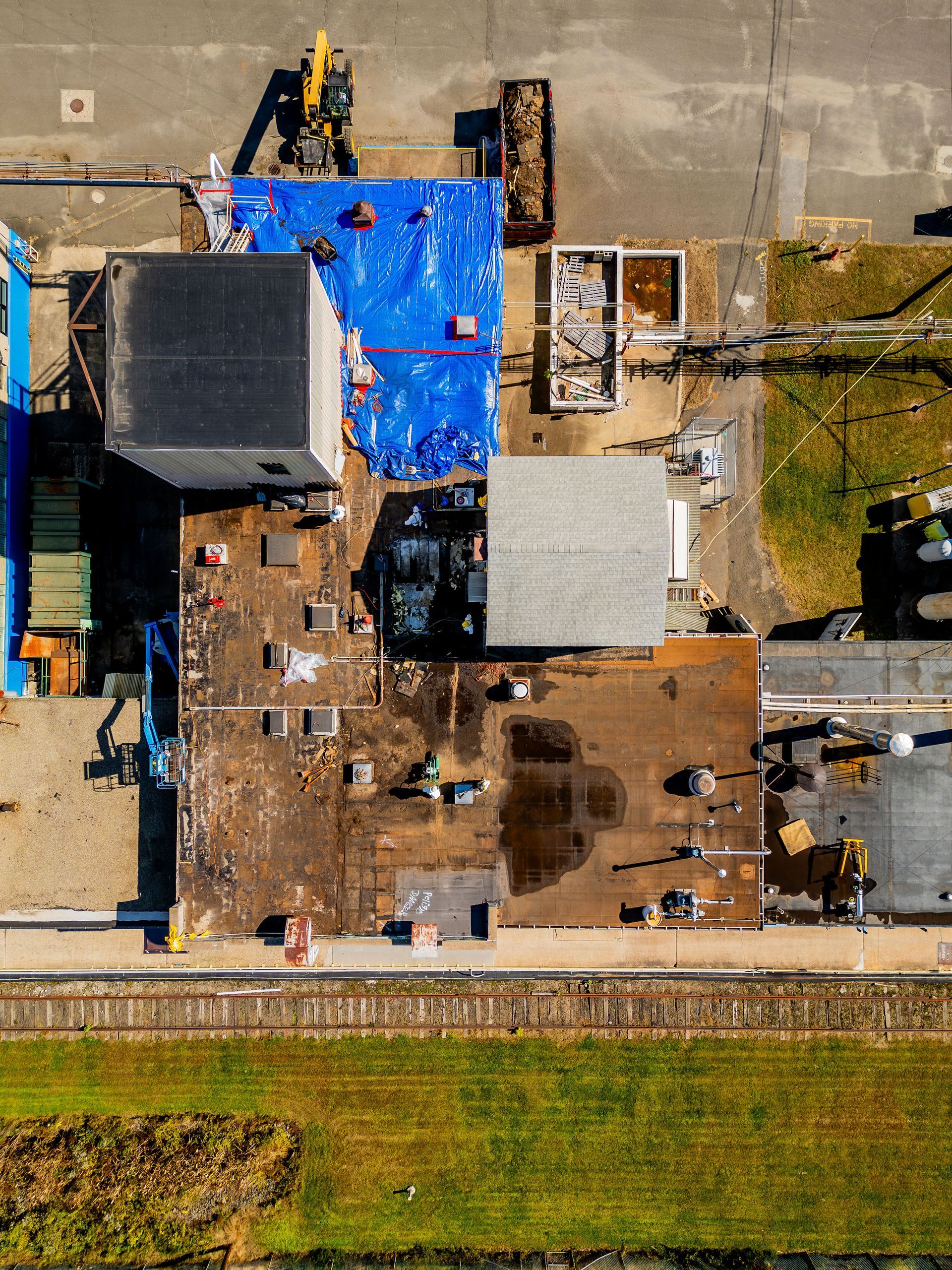 Aerial view of a damaged house with blue tarp sections and a yard beside a road and grassy field