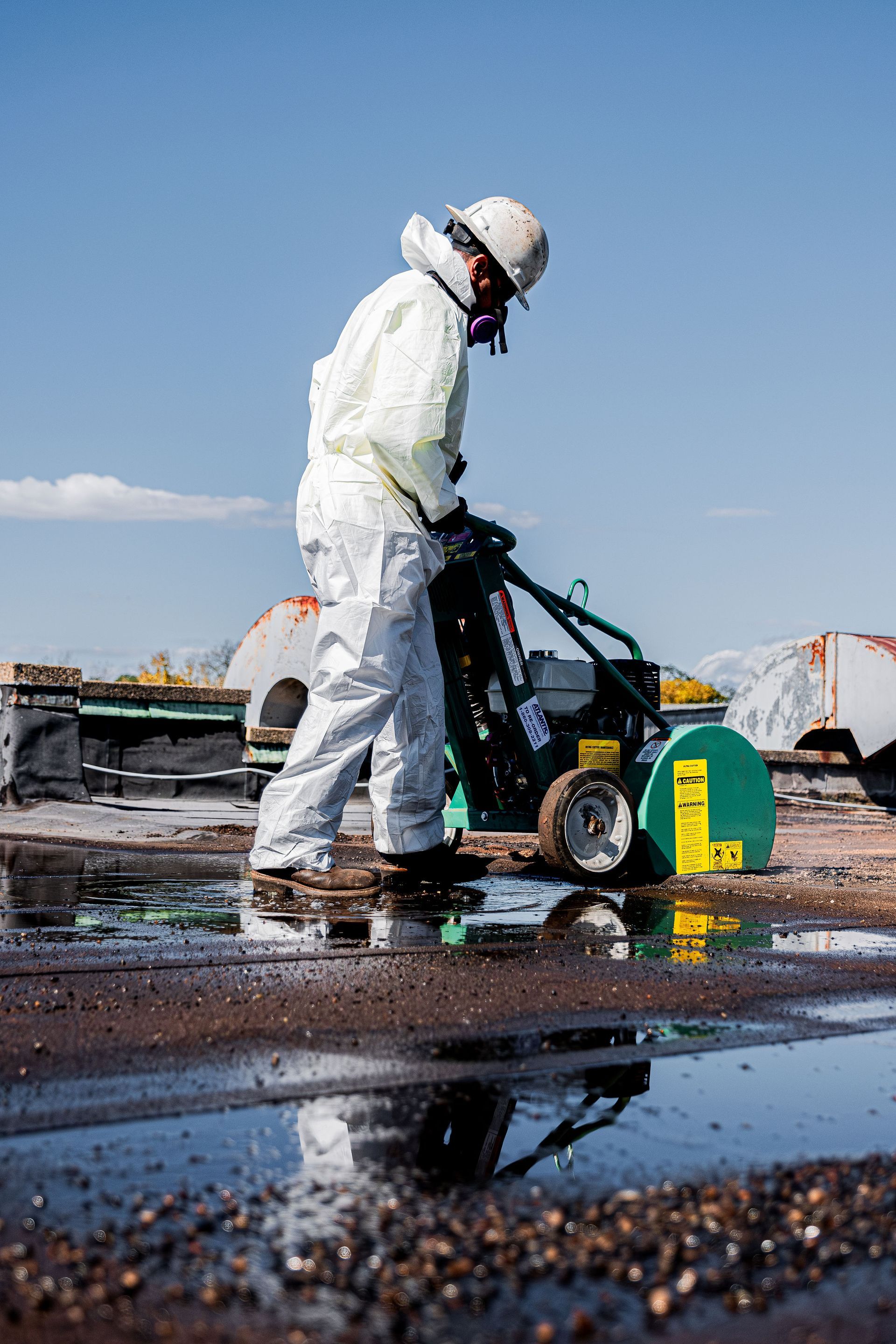 Worker in white protective gear operating a machine on a wet industrial rooftop under a blue sky.