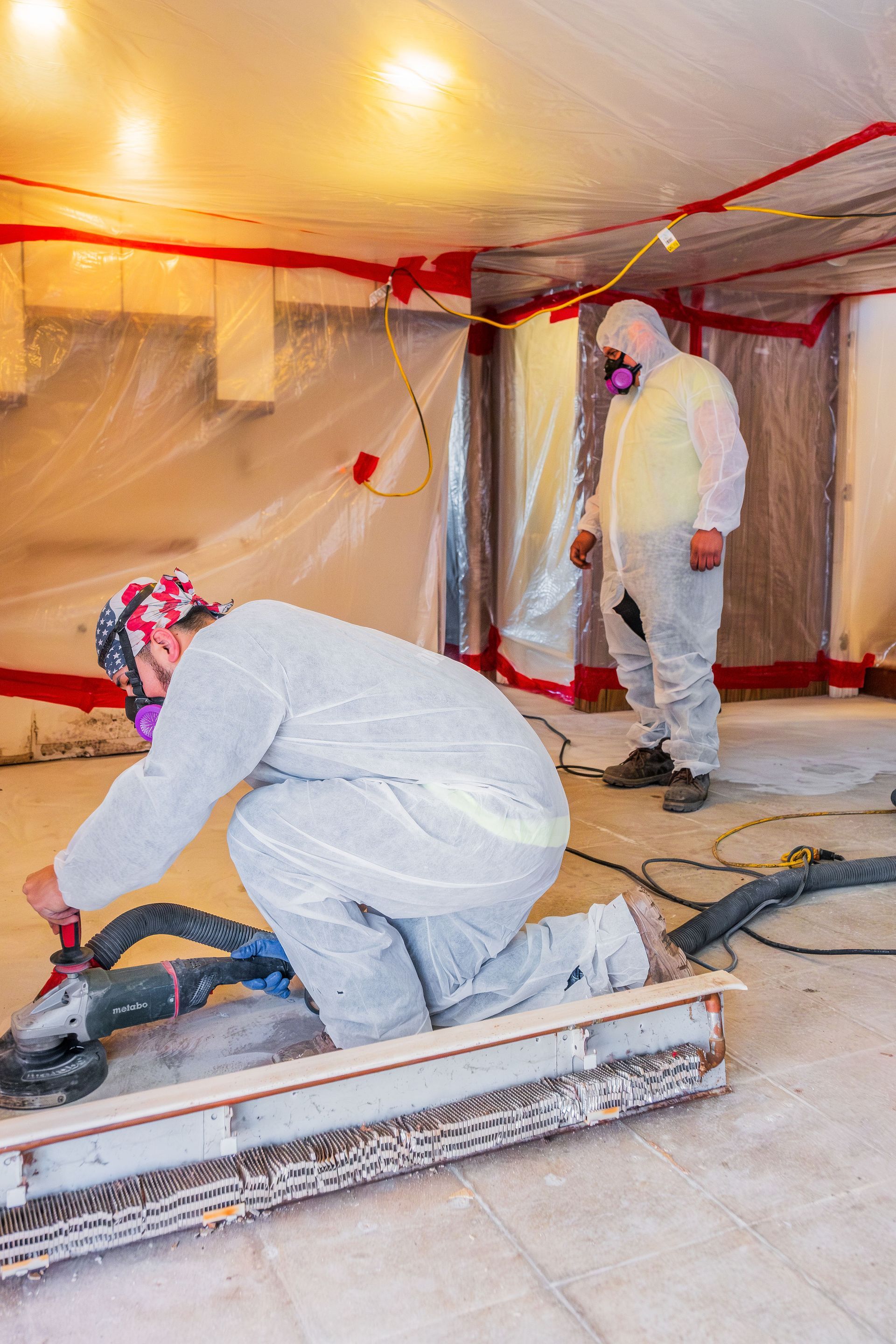 Two workers in white coveralls cutting flooring in a room covered with plastic sheeting and red tape.