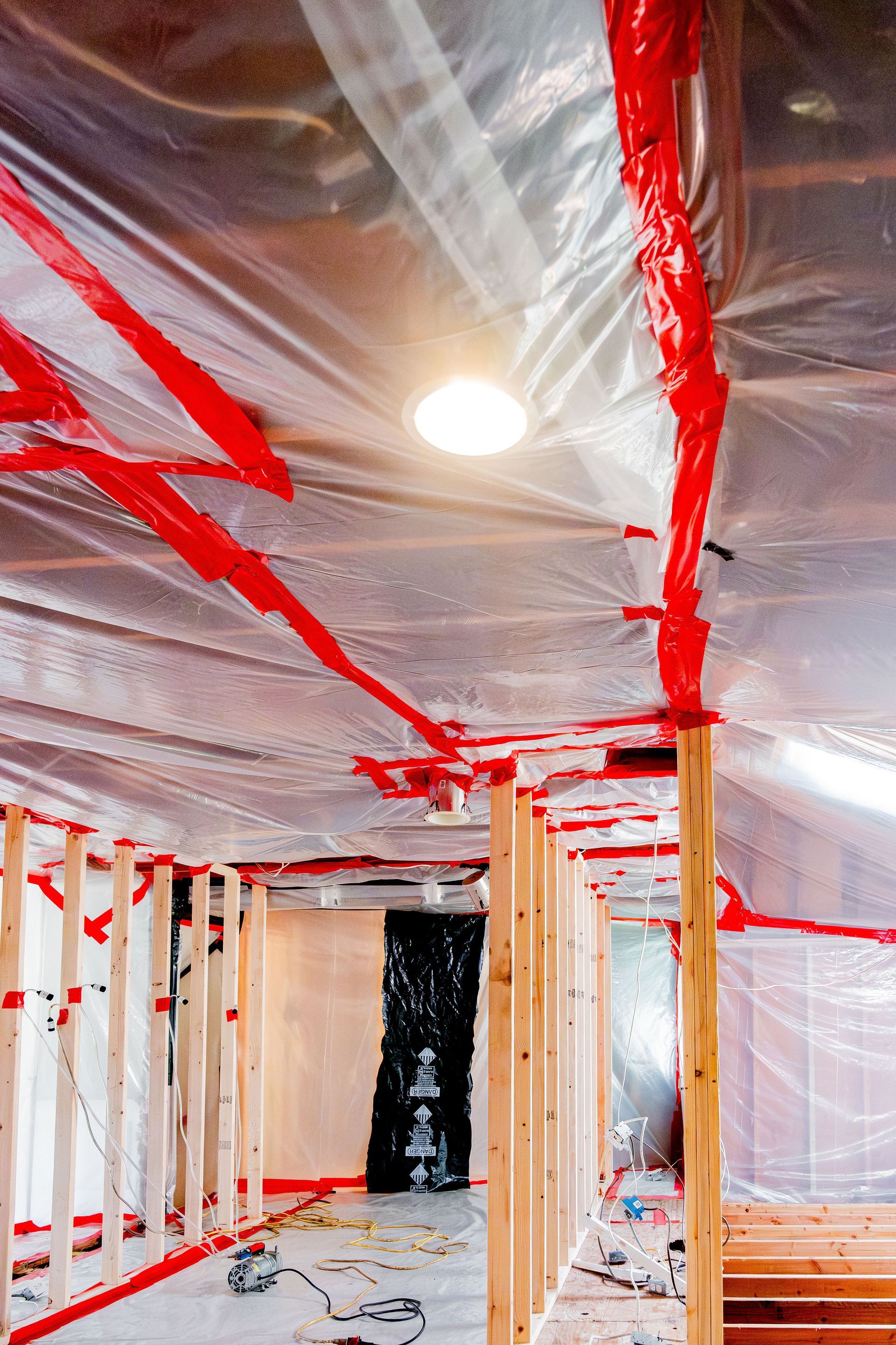 Plastic-covered room under construction with red tape and exposed framing through an unfinished ceiling.