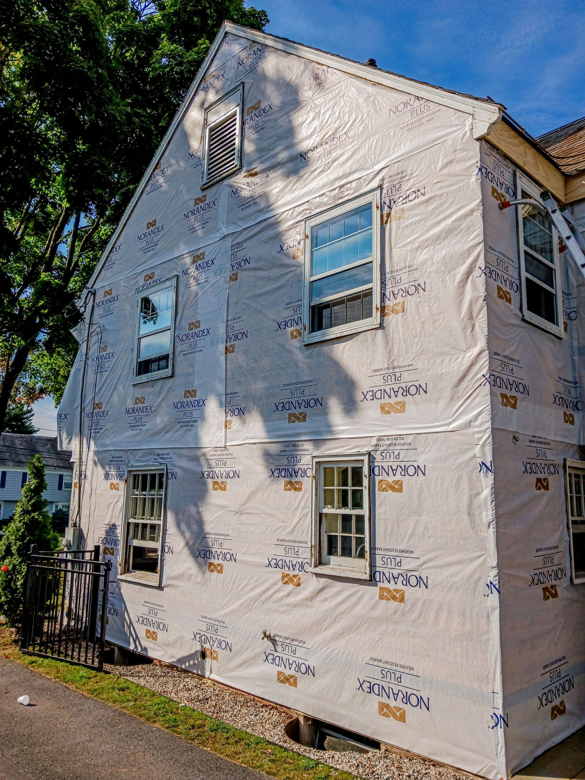 Small two-story house wrapped in weatherproofing, with boarded windows and a tree shading the side.