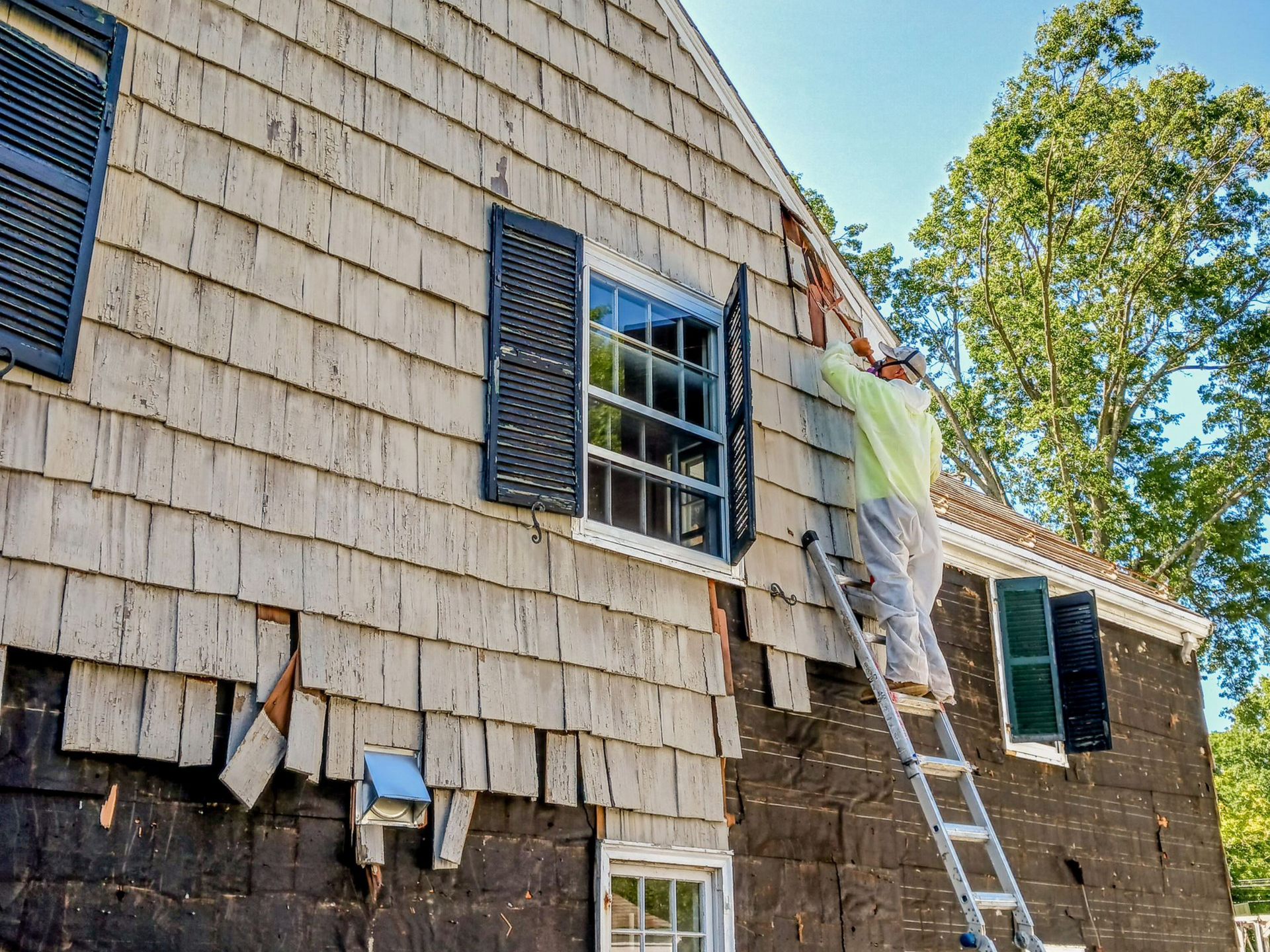 Workers on ladders repairing a shingled house exterior, with dark siding and blue-trimmed windows.