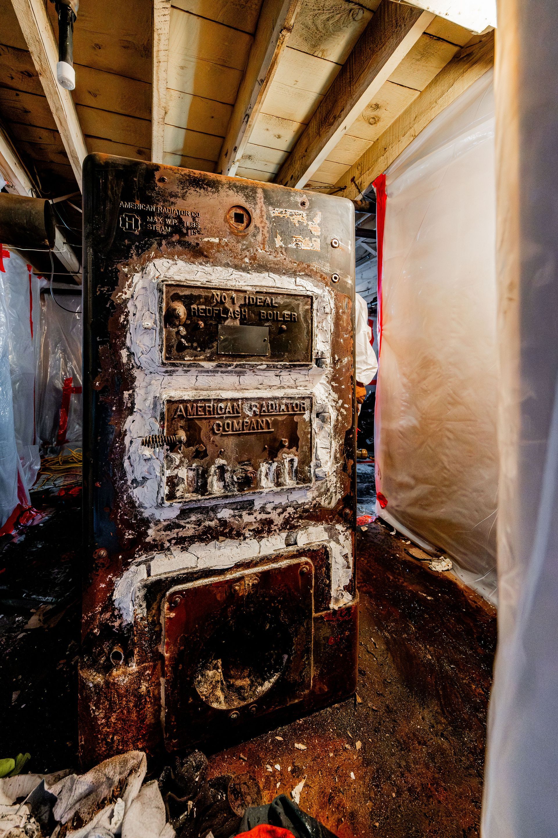 Rusty furnace in a basement utility room with exposed beams and plastic sheeting.