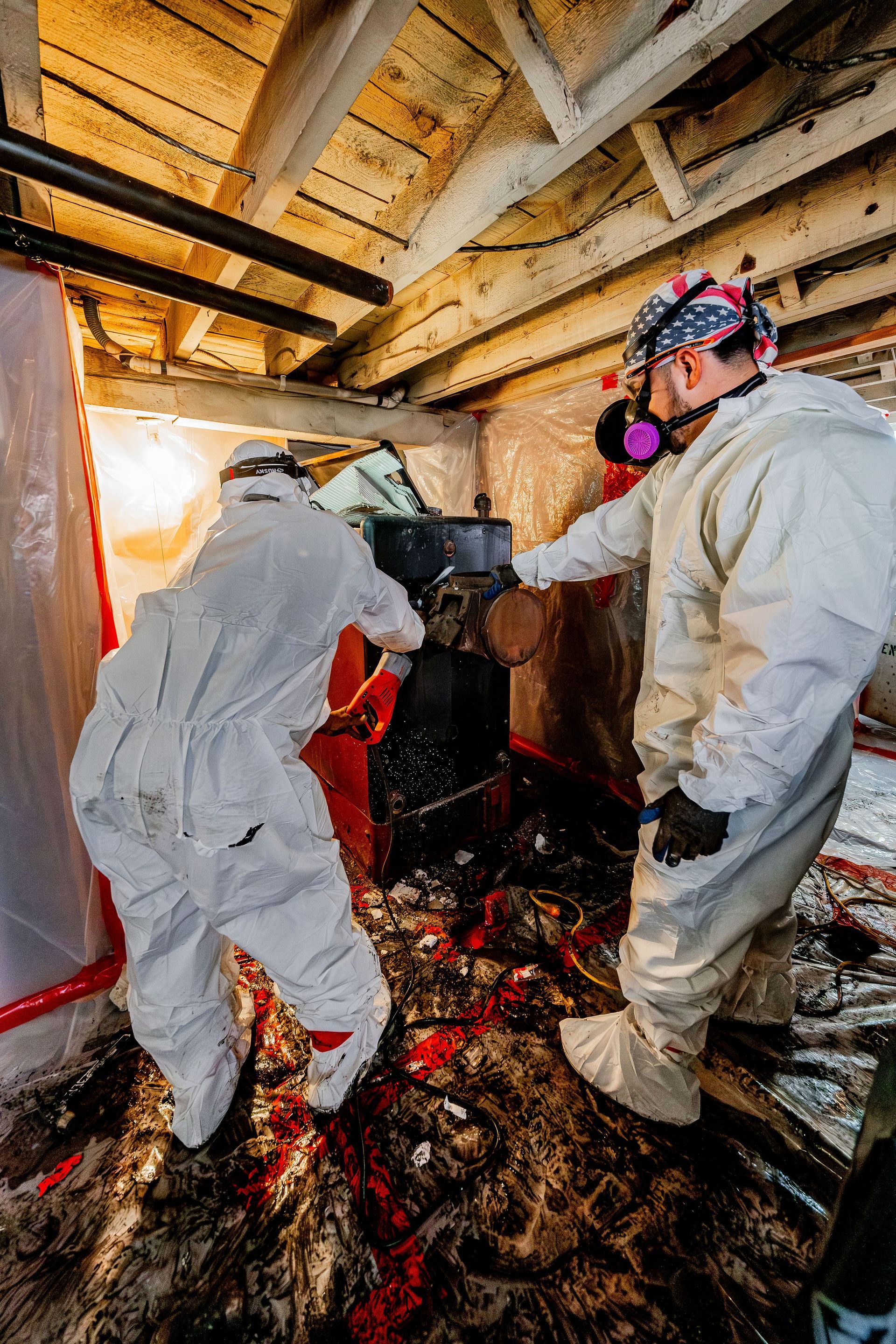 Two workers in protective suits and masks inspect a fire-damaged room with debris on the floor.