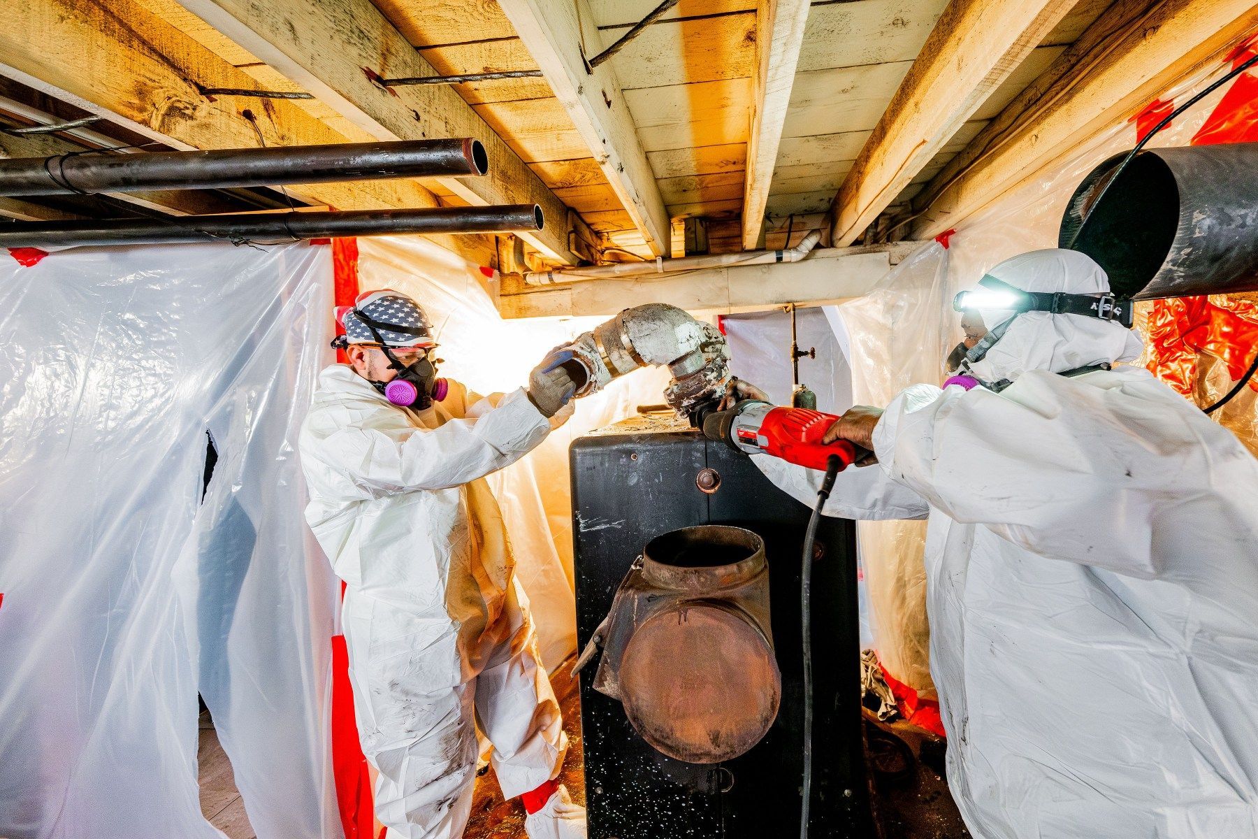 Two workers in white protective suits handling equipment in a basement with exposed beams and pipes.
