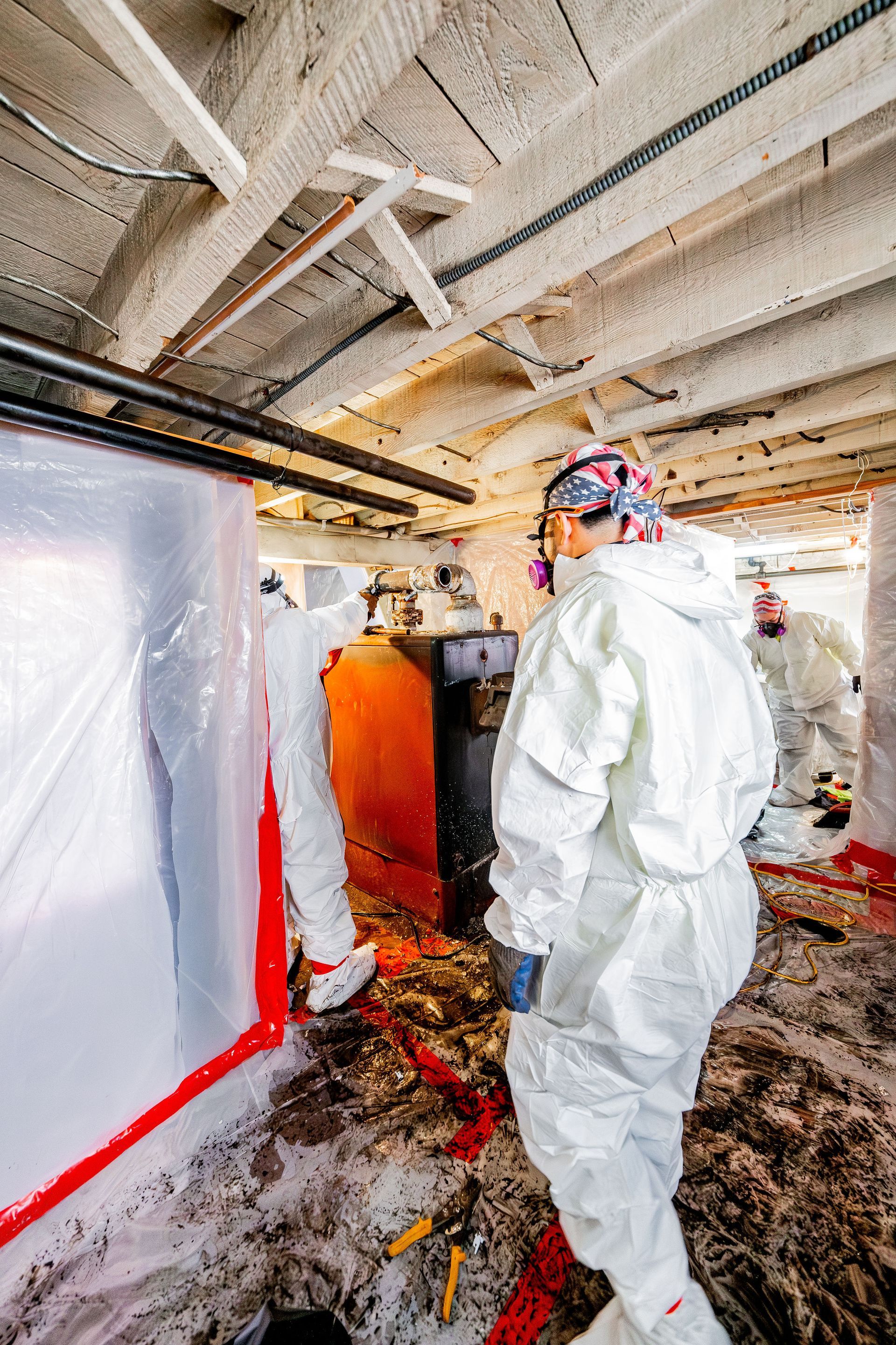 Workers in protective suits inspecting an industrial ceiling and equipment in a damaged, debris-filled room