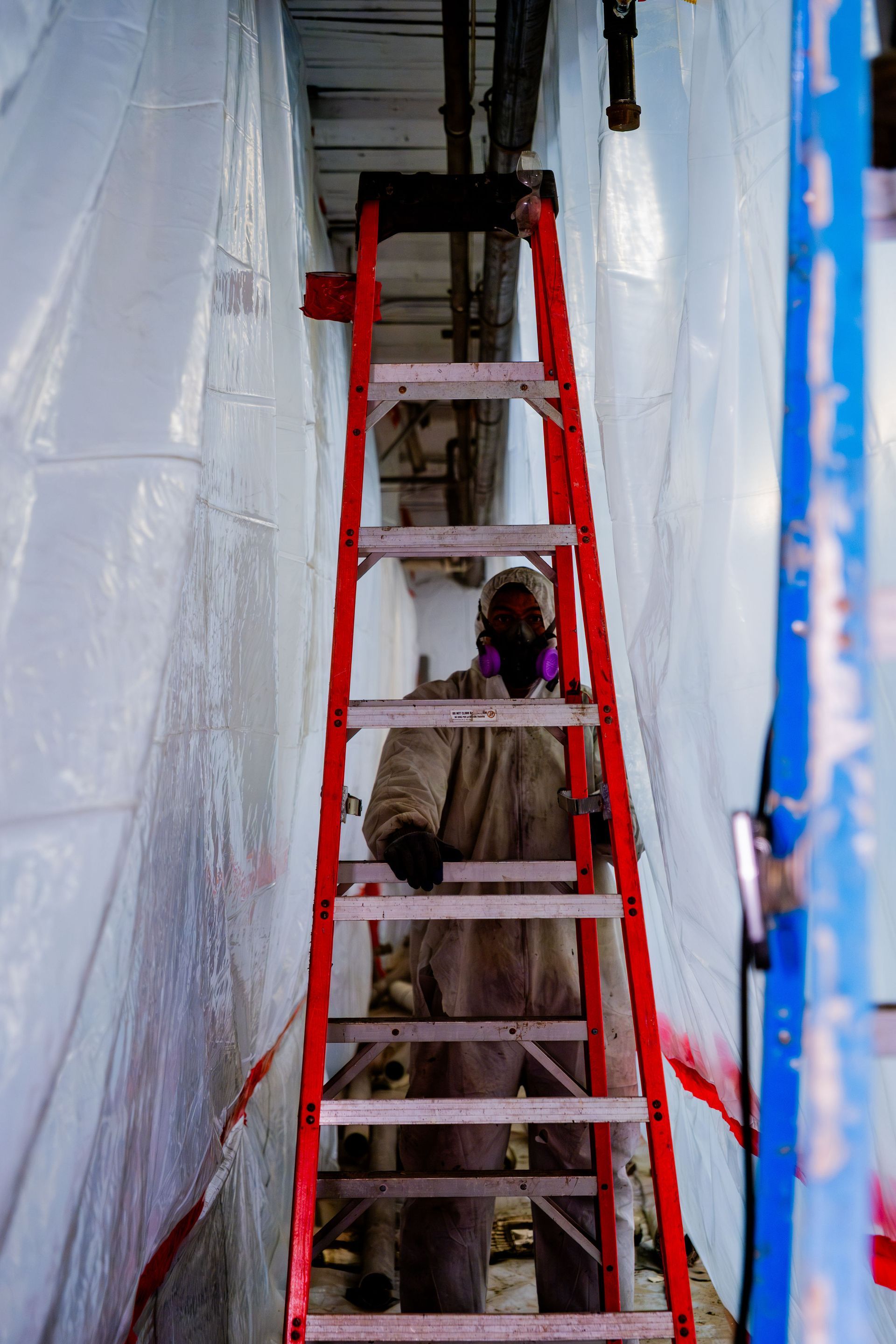 Person on a ladder in a narrow taped-off corridor, surrounded by white plastic sheeting and blue trim