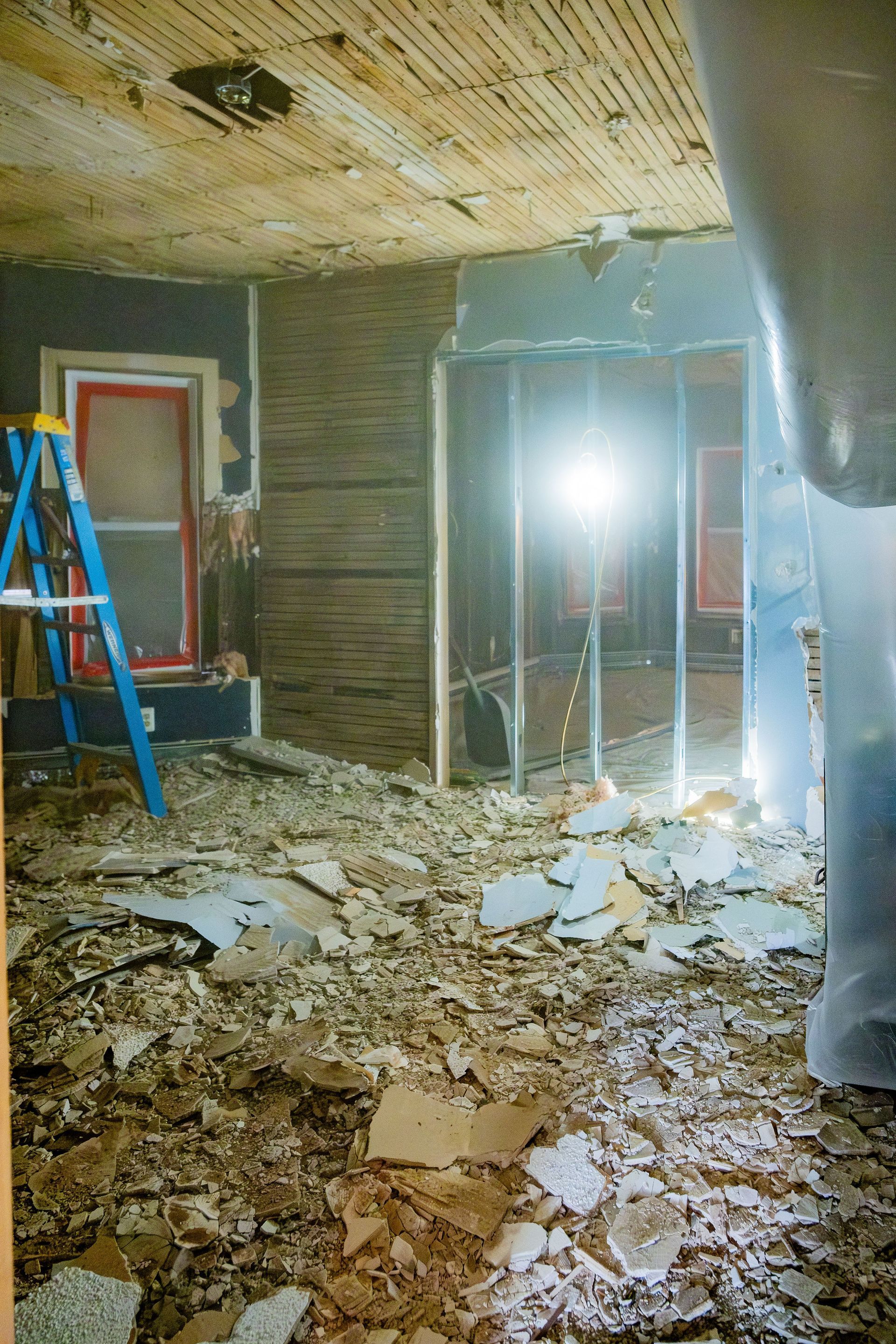 Damaged room with debris-covered floor, exposed walls, and a blue ladder under renovation.