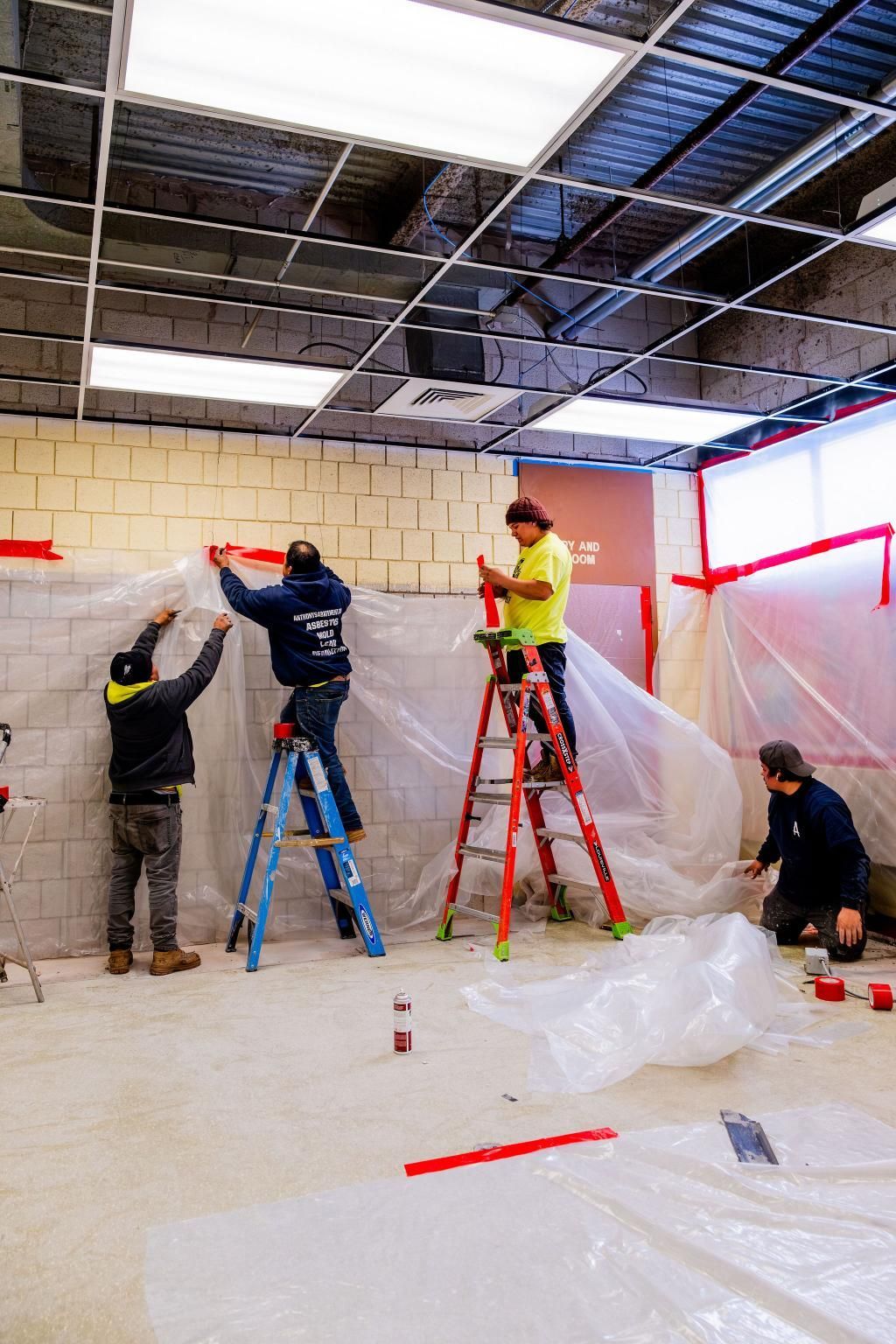 Workers installing ceiling panels in a room, some on ladders, with plastic sheeting covering the floor.