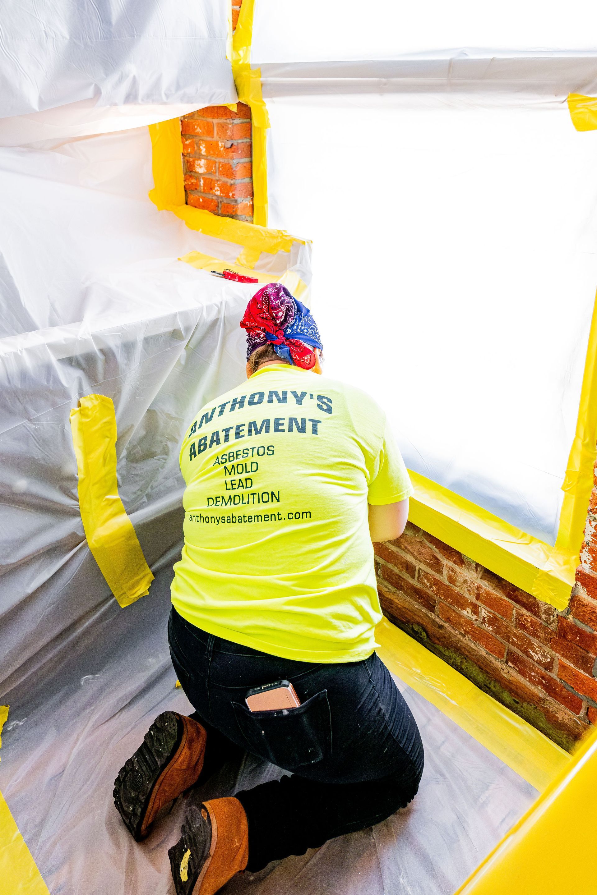 Person kneeling in a room covered with plastic sheeting, painting a wall white with a brush.