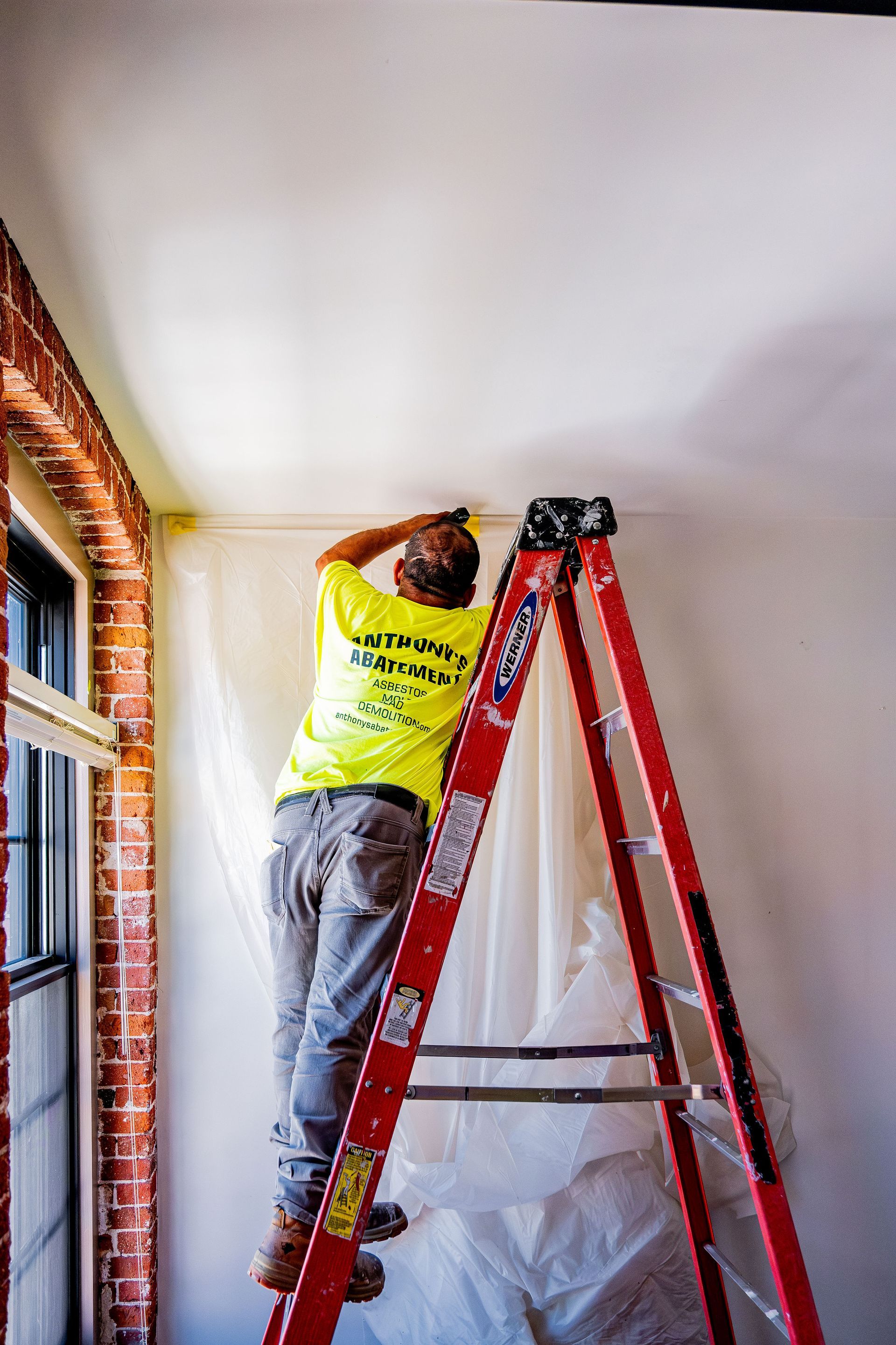 Worker on a red ladder painting a white ceiling in a room with large windows